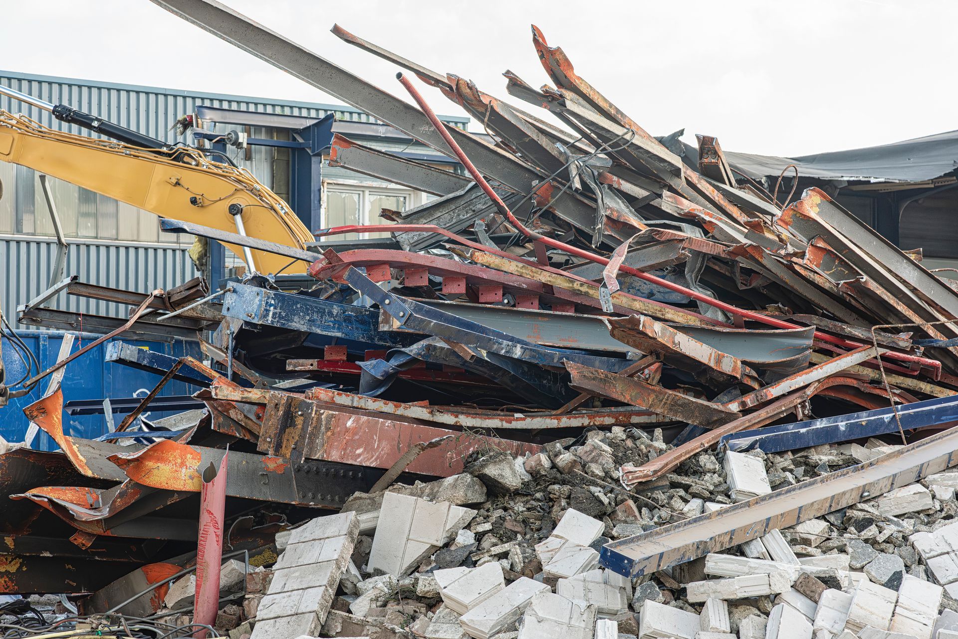 Demolished building rubble with an excavator arm; mostly blue, red, and silver metal with white concrete debris.