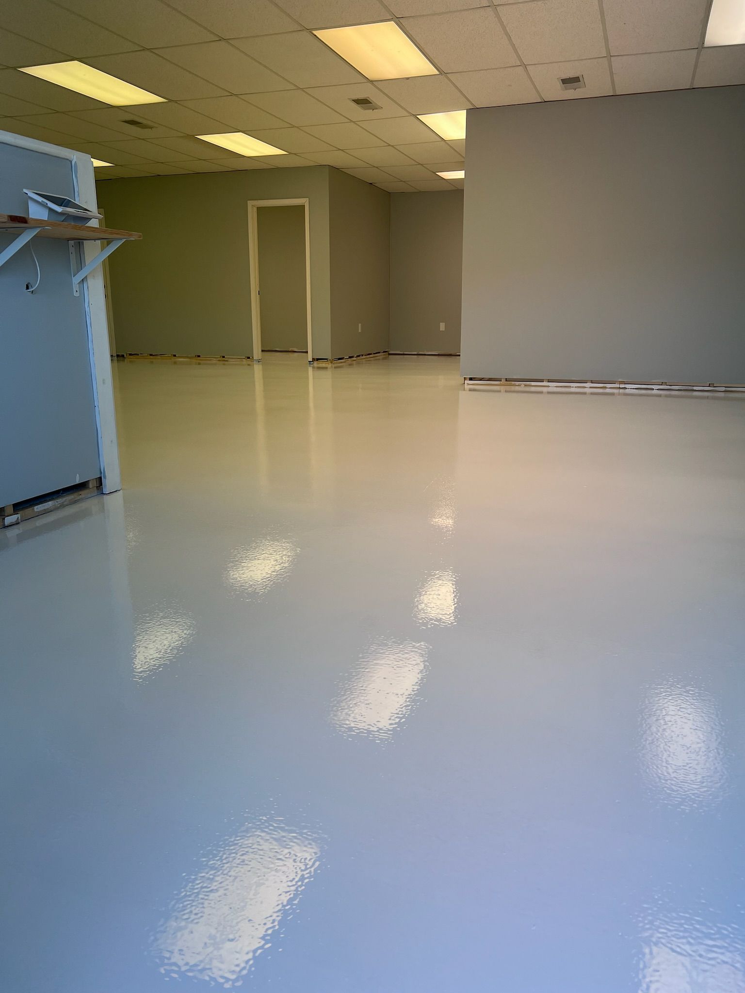 Empty room with glossy, light-colored floor reflecting overhead lights. Gray walls and a doorway are visible.