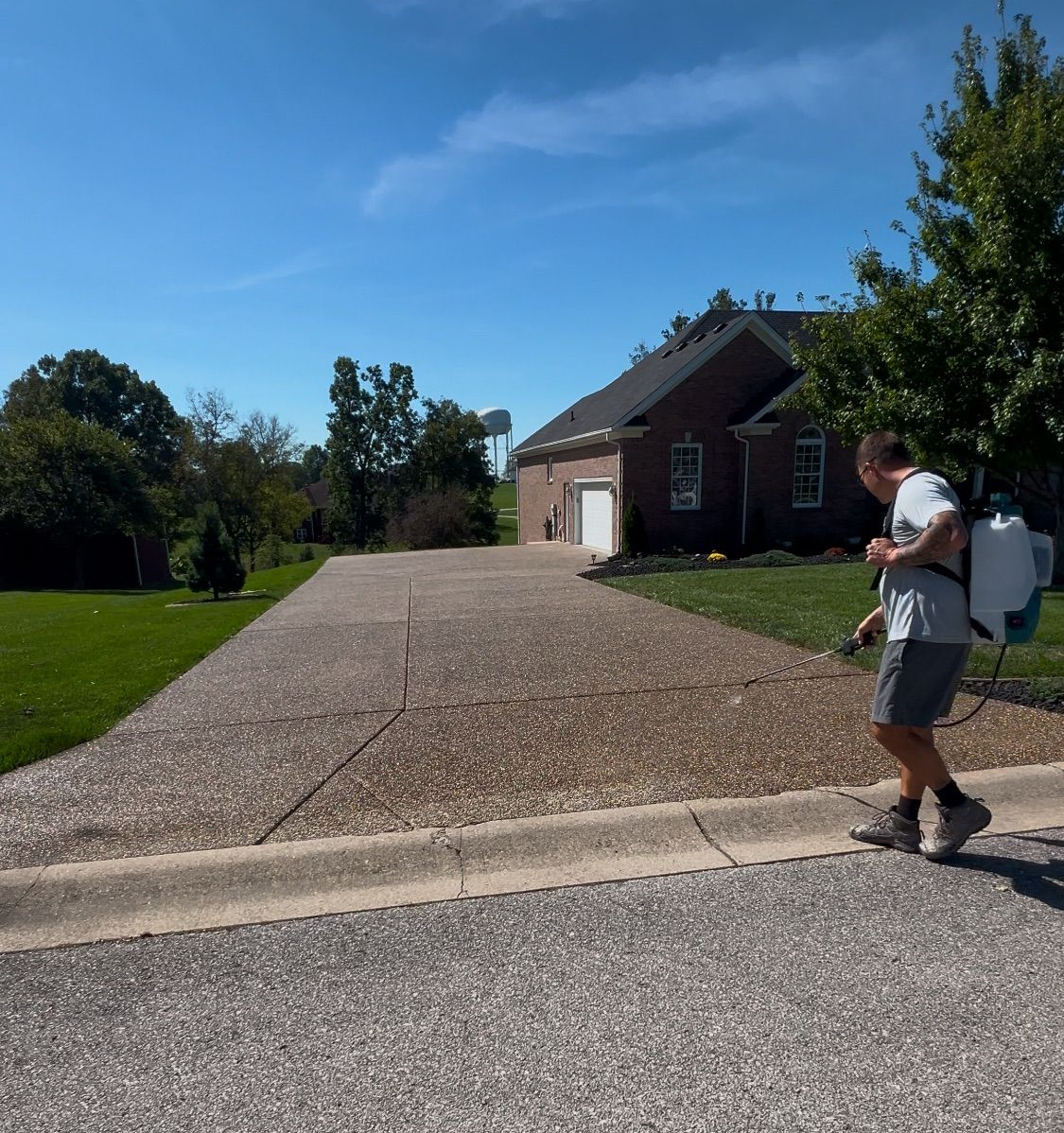 Man spraying driveway with a backpack sprayer on a sunny day in front of a brick house.