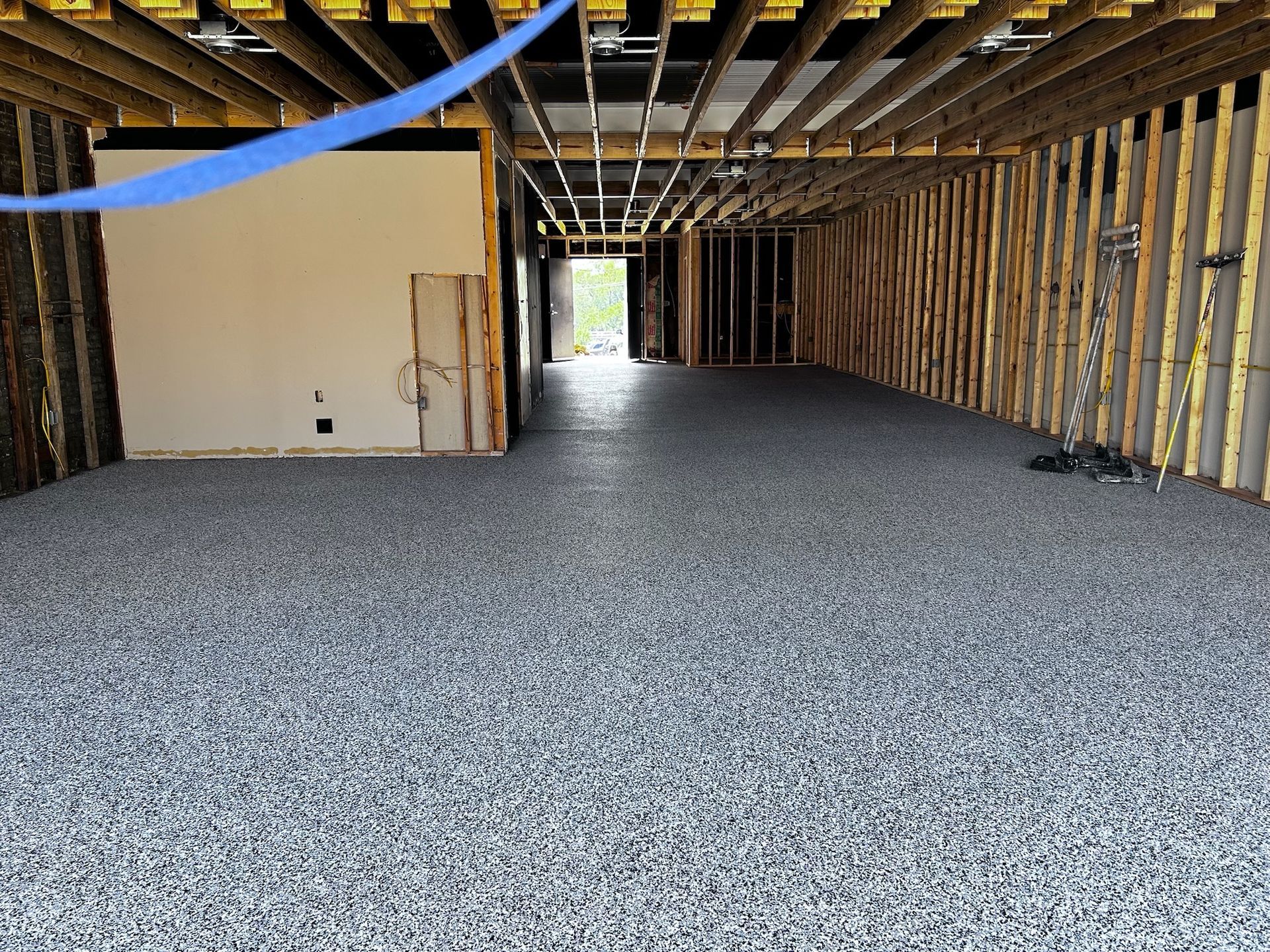 Interior of a building under construction, showing a gray, granular flooring and exposed wooden beams and studs.