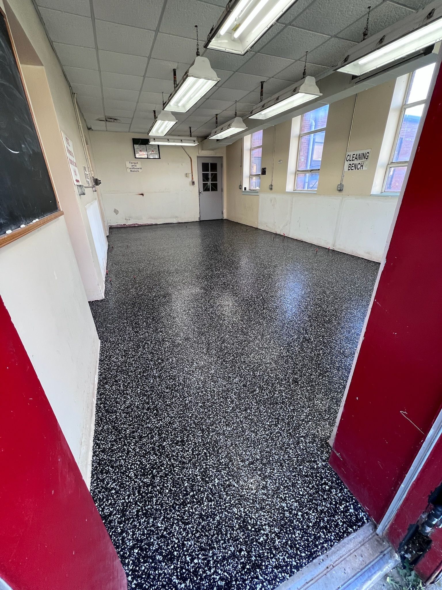 Empty room with speckled black and white floor, fluorescent lights, and windows.