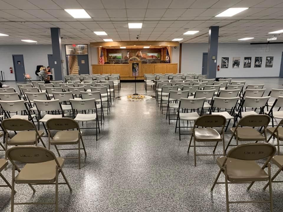 Rows of beige folding chairs face a stage in a large hall with a speckled floor and neutral walls.