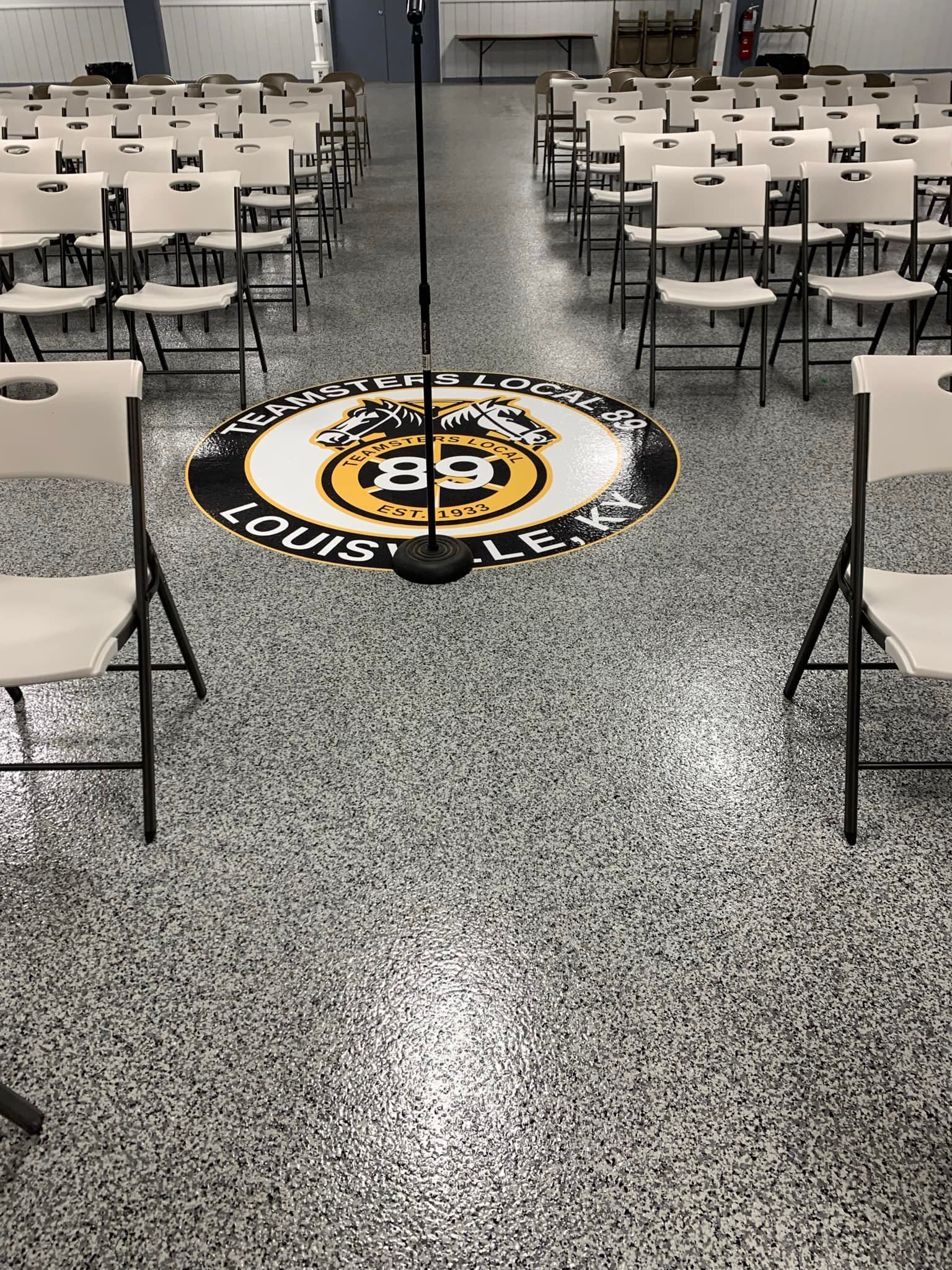 Empty room with rows of chairs, microphone on stage with logo, speckled floor.