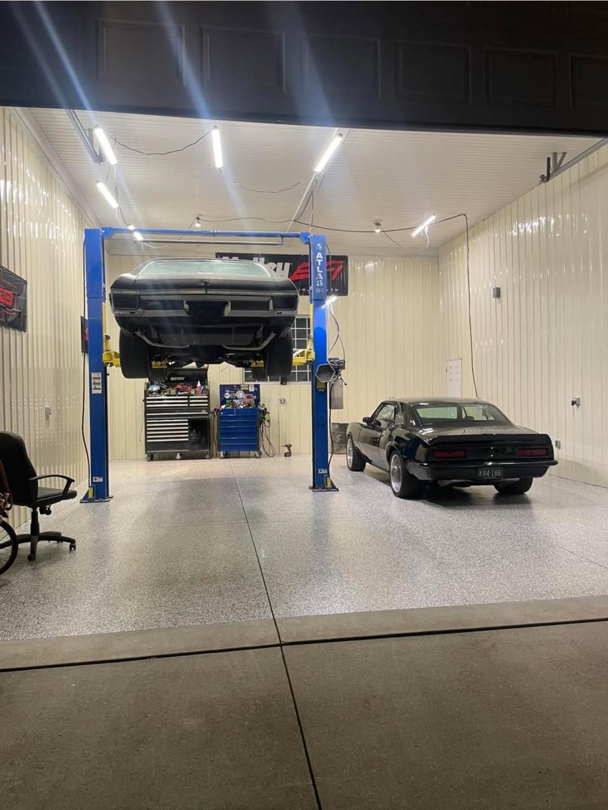 Two classic cars in a car repair shop. One on a lift, one on the floor. Shop has a gray speckled floor, and white walls.