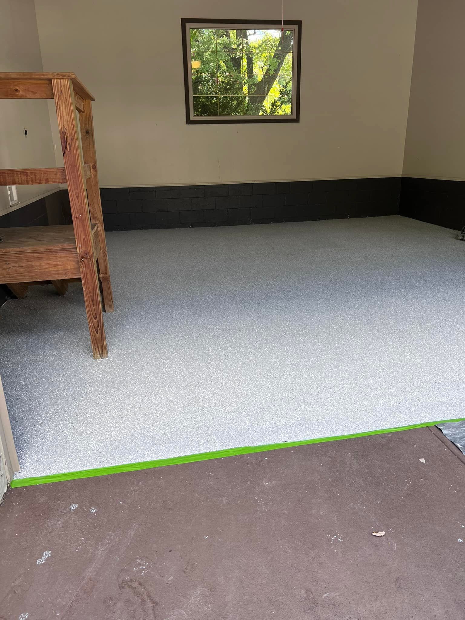 Interior of a room with a light gray speckled floor. A window is in the back wall. A wooden ladder is on the left.