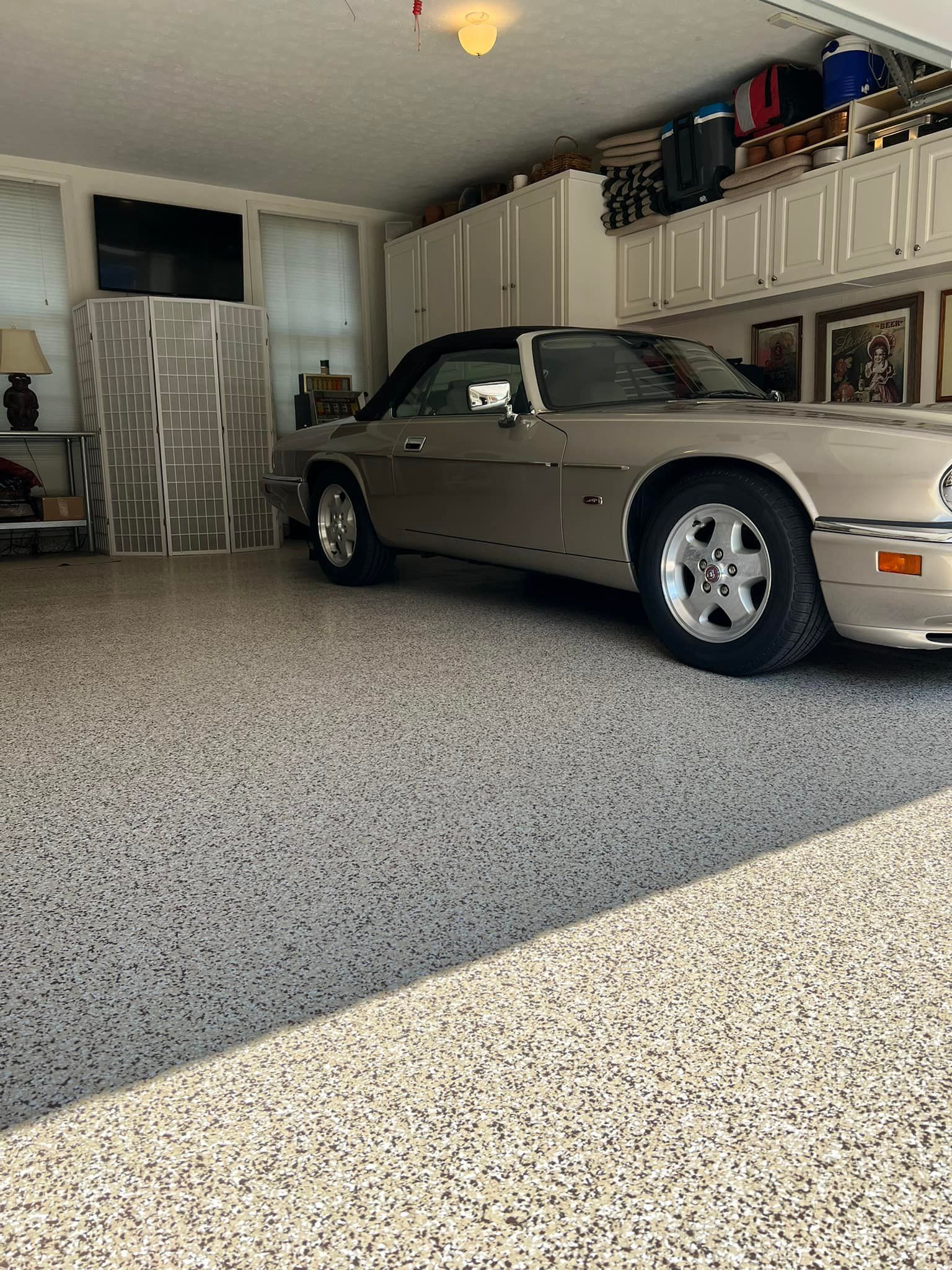 Tan Jaguar convertible in a garage with a speckled floor. Cabinets and a television are visible.