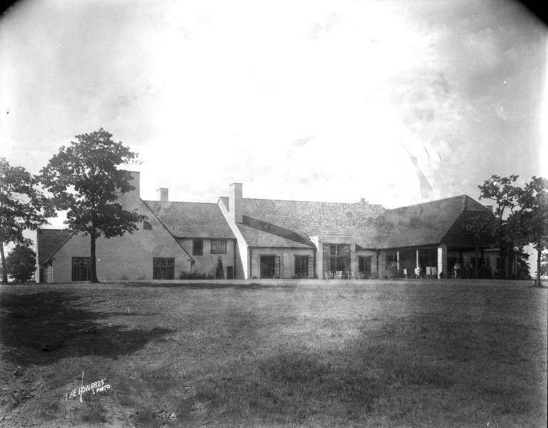 Black and white photo of a large house with a long, low roof, on a grassy lawn under a cloudy sky.
