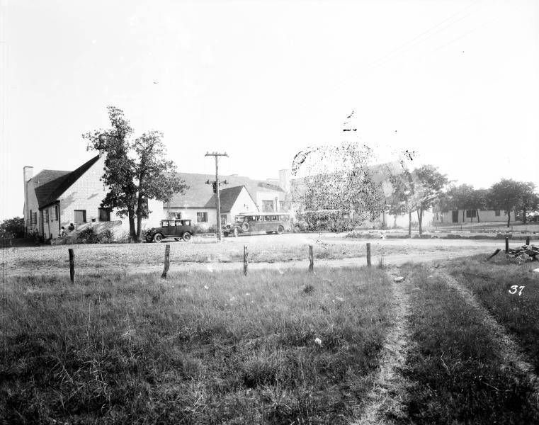 A black and white photo shows a grassy field with a path leading to buildings and power lines in the distance.