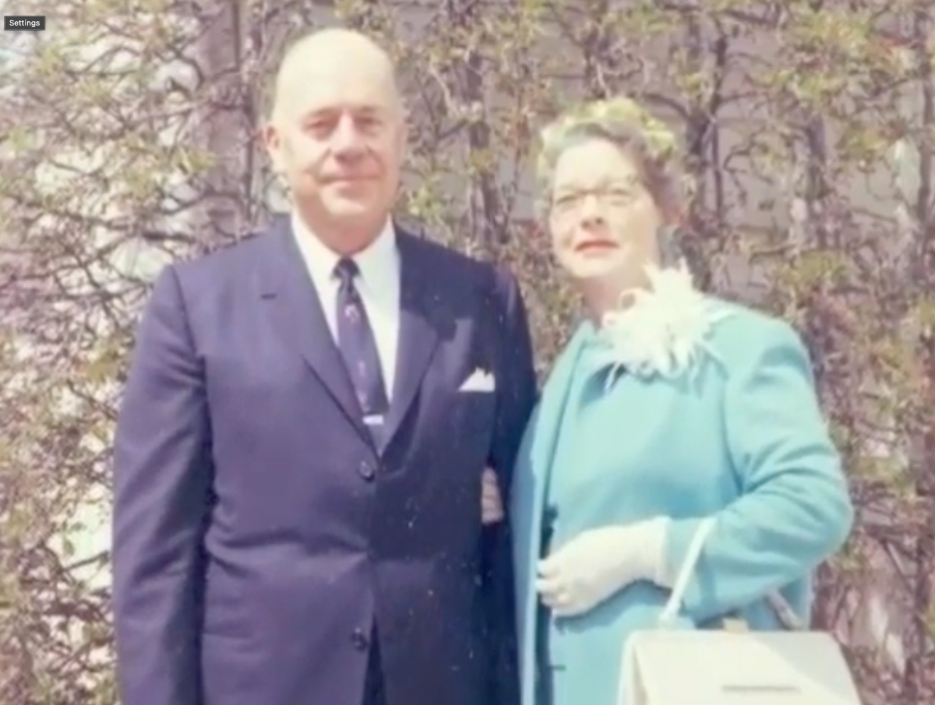 Man and woman in formal attire pose outside. Man in navy suit, woman in blue coat, holding a purse.
