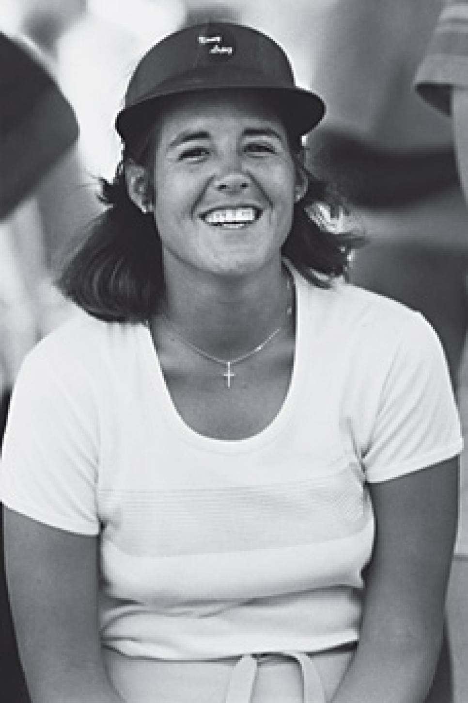 Woman wearing a cap and necklace smiles broadly, wearing a white t-shirt.