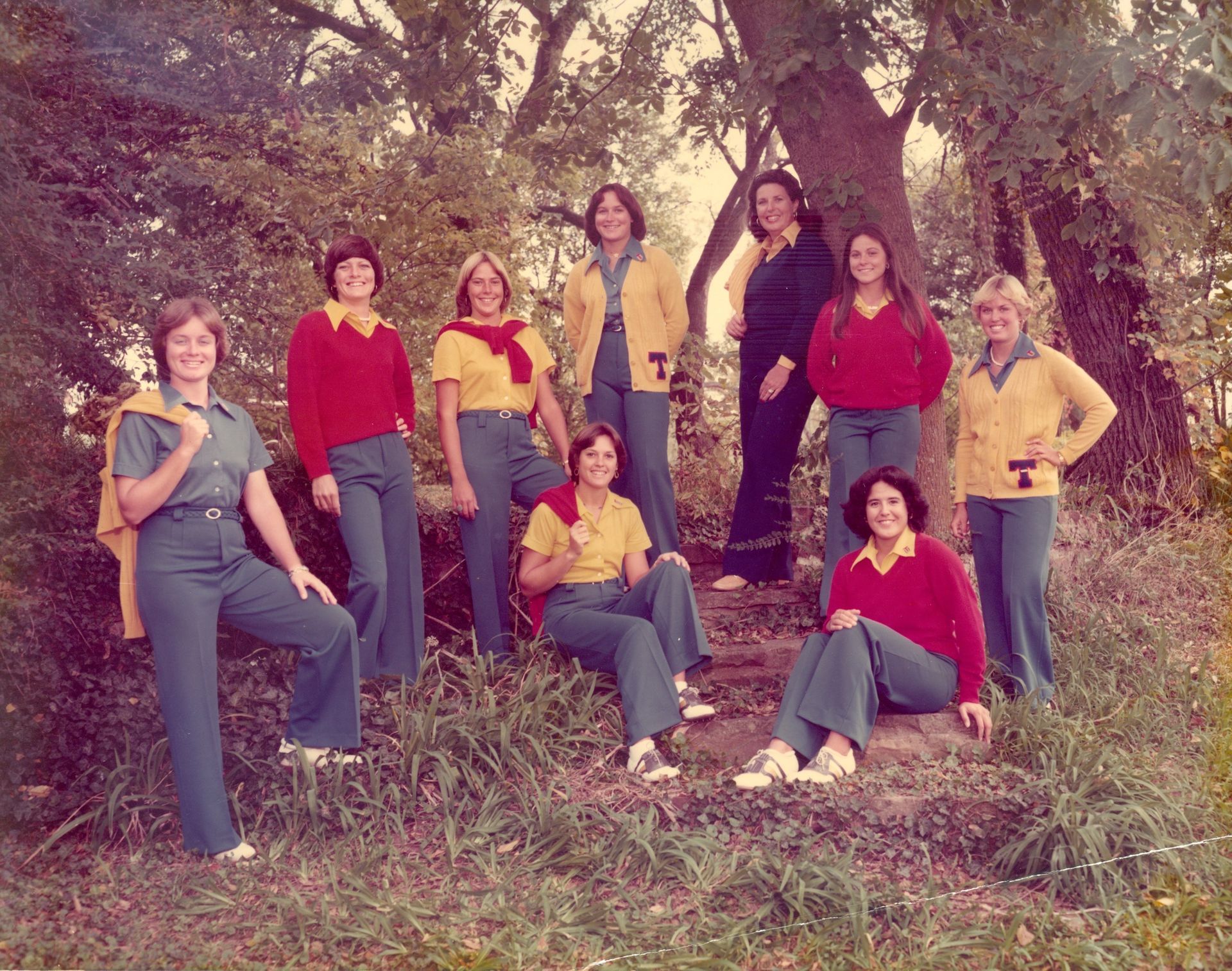 Group of women in yellow sweaters, red shirts, and blue jeans posing outdoors near a tree.