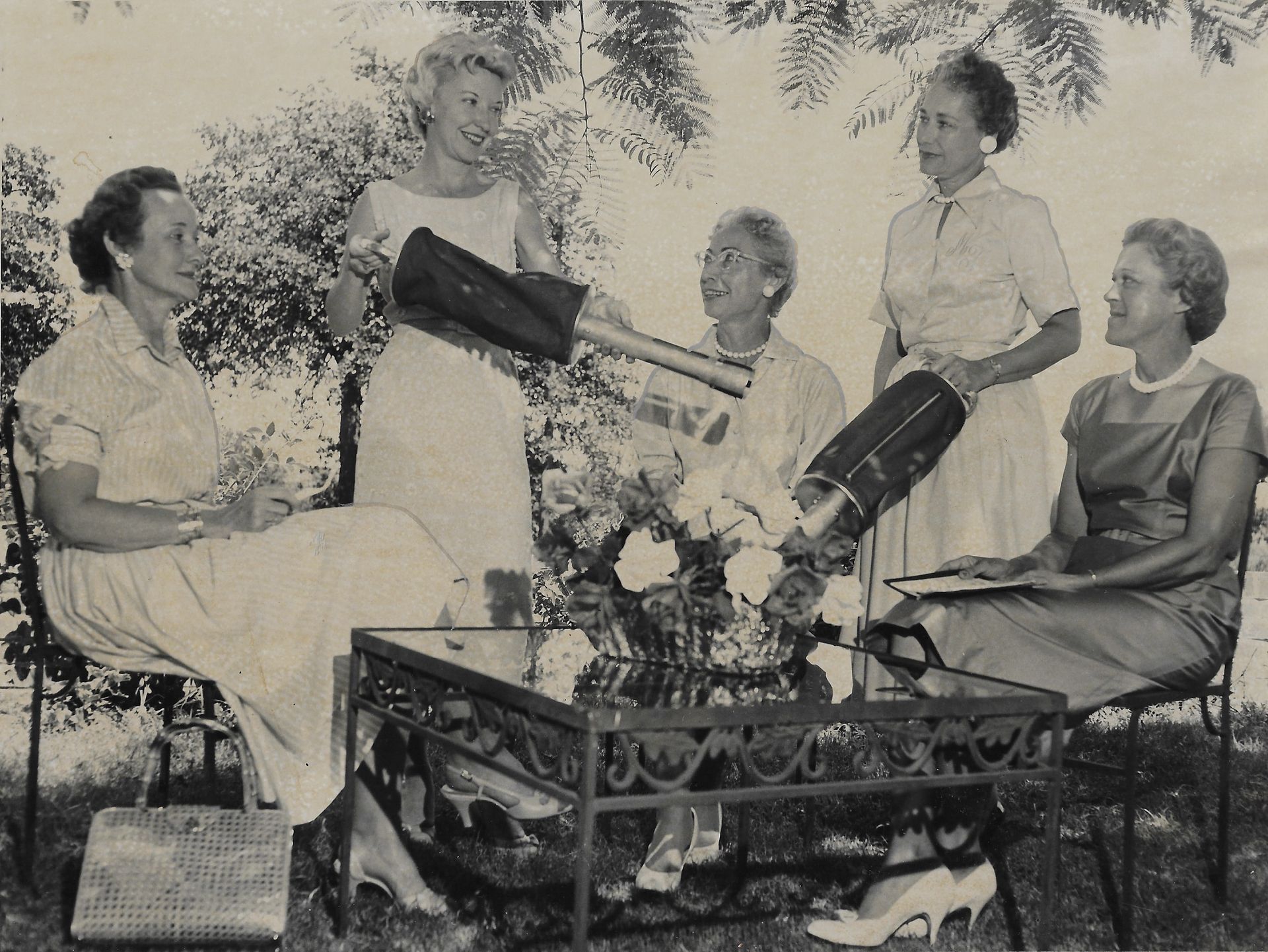 Five women outdoors, one holding a golf bag, gathered around a glass-topped table with flowers.