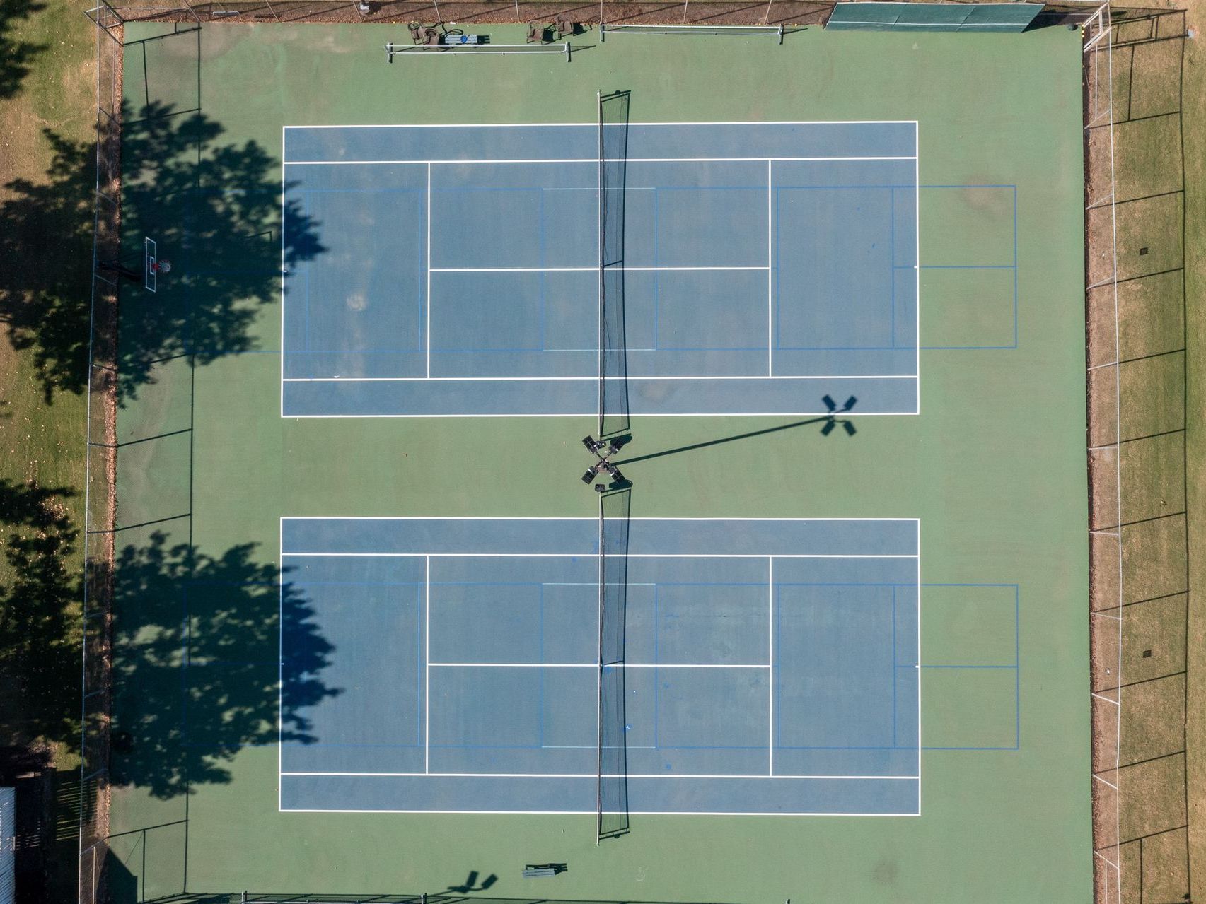 Overhead view of two blue tennis courts, separated by a net, surrounded by green space.