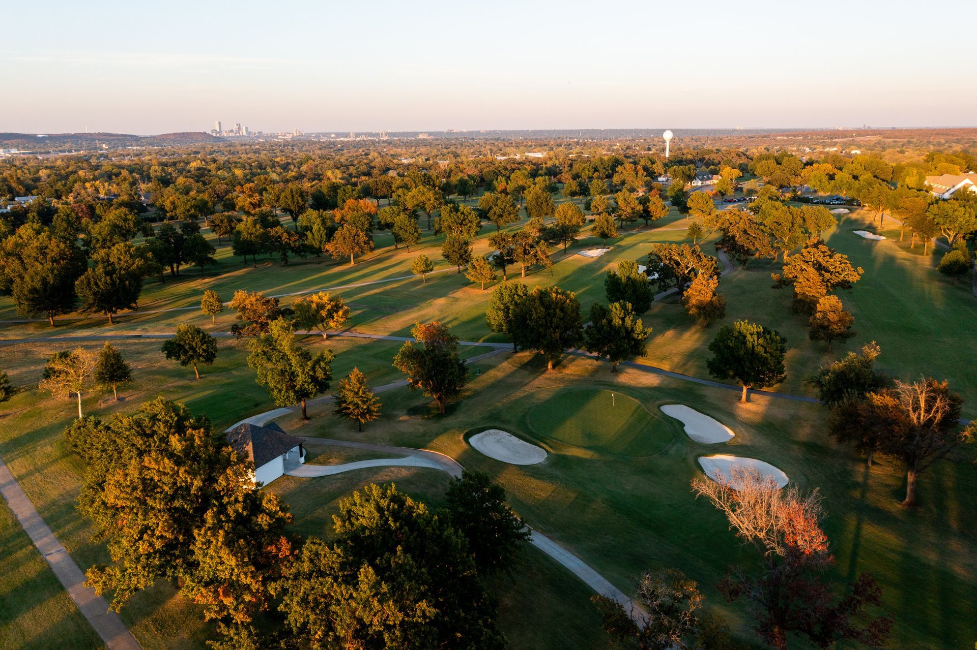 Aerial view of a green golf course with trees, bunkers, and a small building under a warm sunset sky.