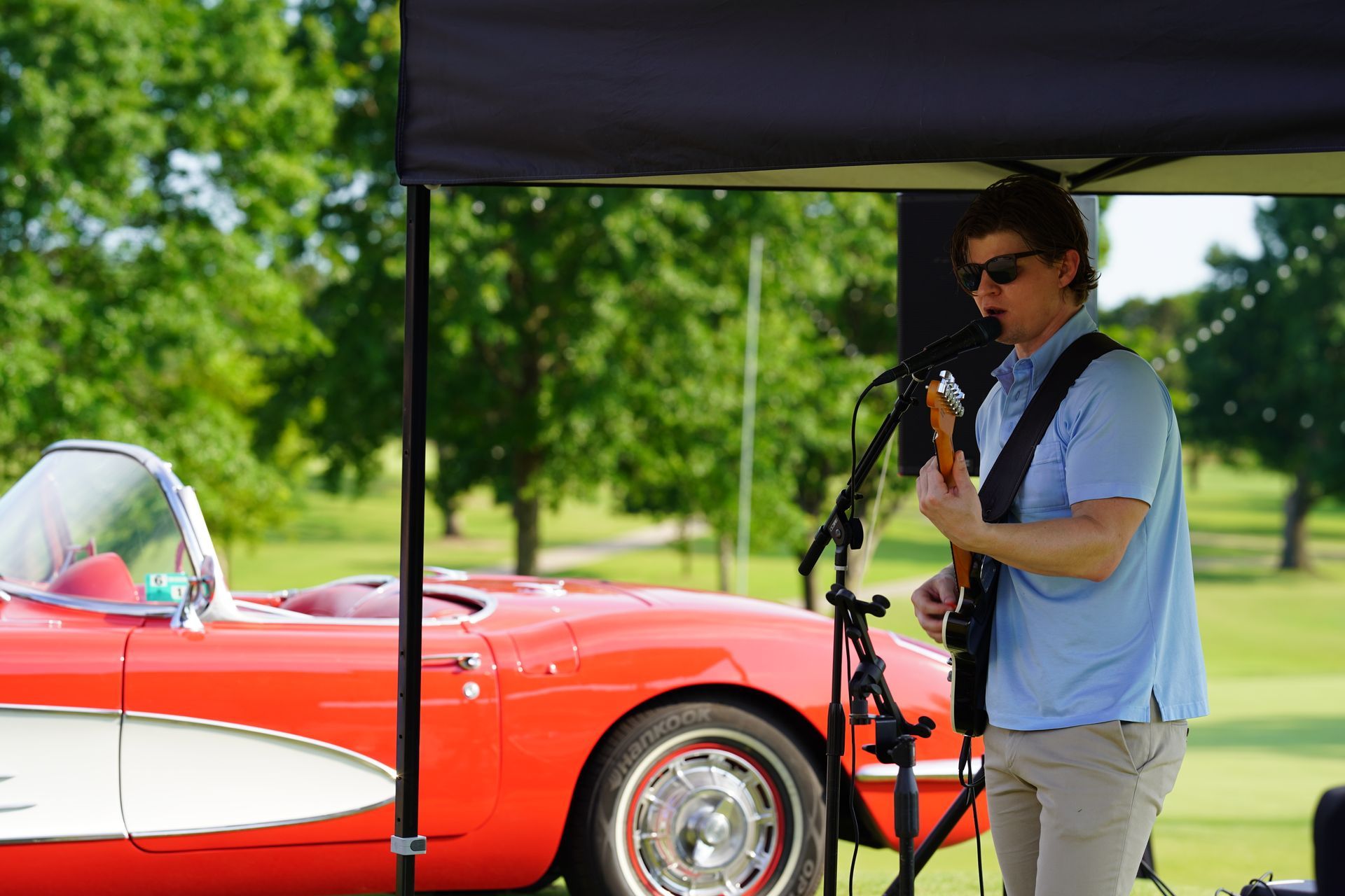 A musician plays guitar under a canopy next to a classic red convertible car on a golf course.