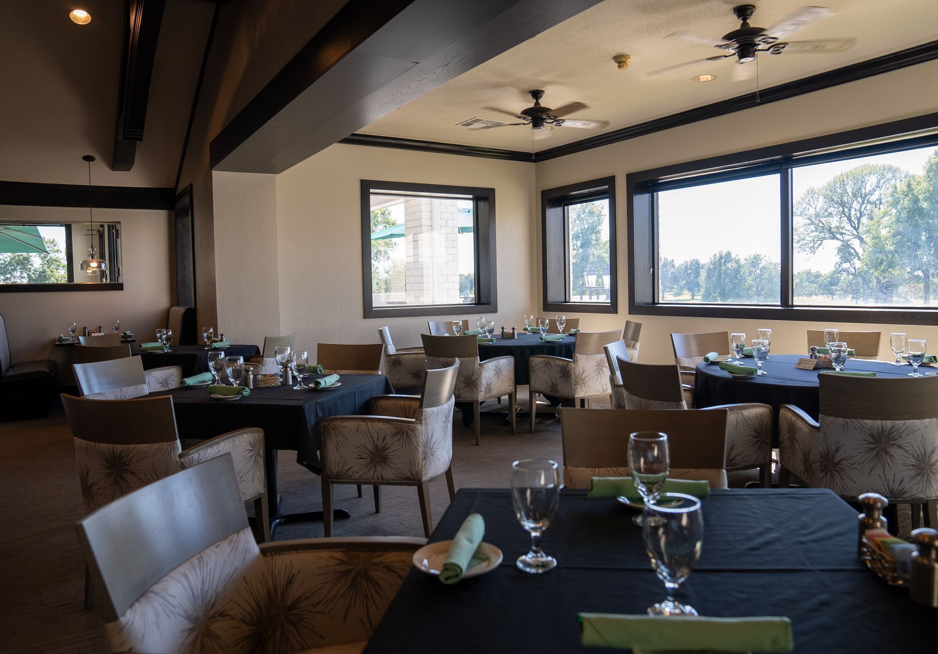 Restaurant dining room with tables, chairs, windows, and ceiling fans. Tables have black tablecloths and place settings.