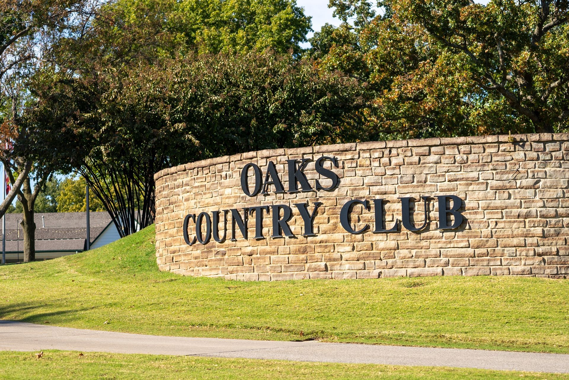Sign for Oaks Country Club on a stone wall, text in black, with green grass and trees in the background.