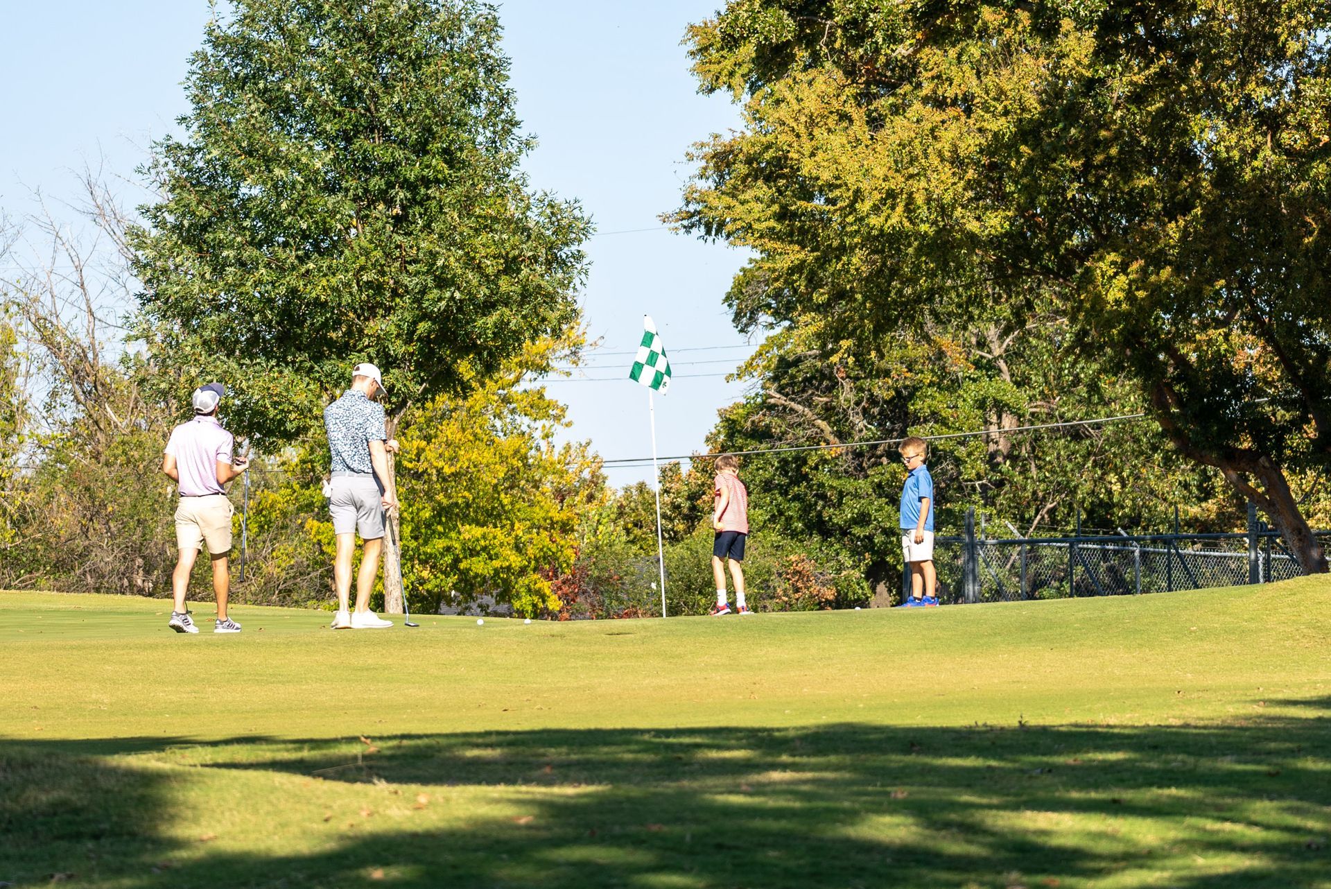 Four people on a golf course. Trees frame the green with a flag. Sunny day.