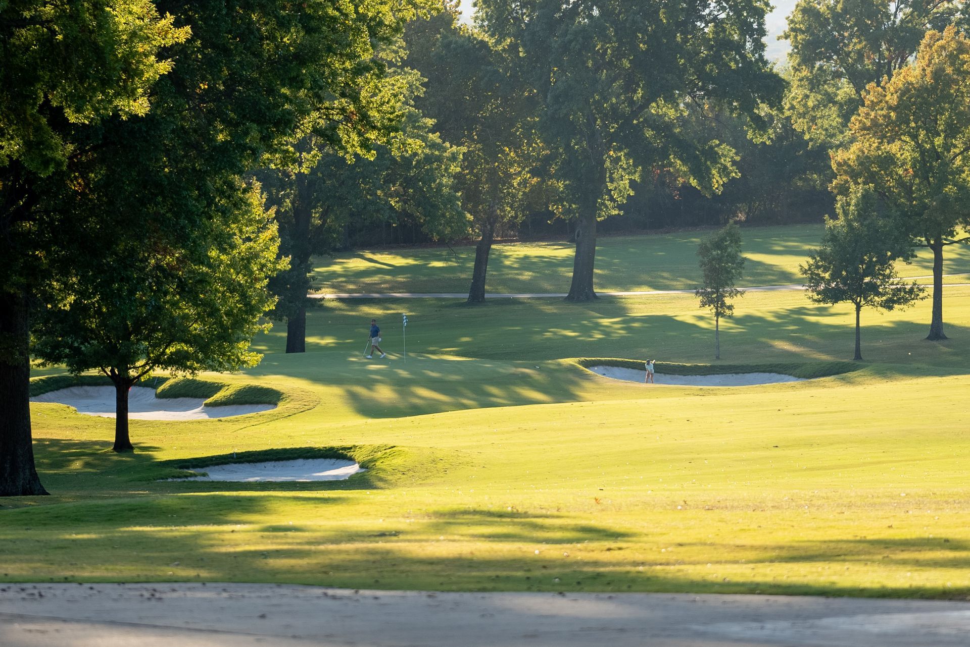 Golf course scene with green grass, trees, and sand traps under sunlight.