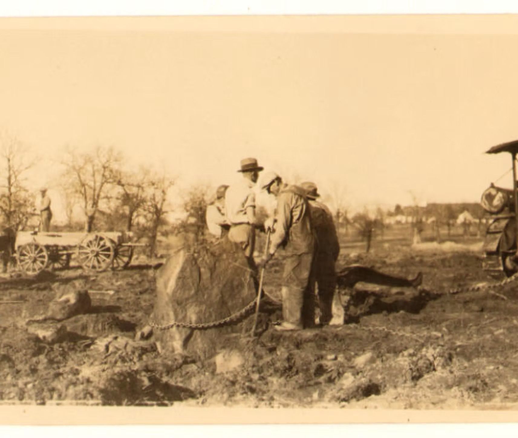 Men moving a large boulder in a field, with a cart and machinery visible.