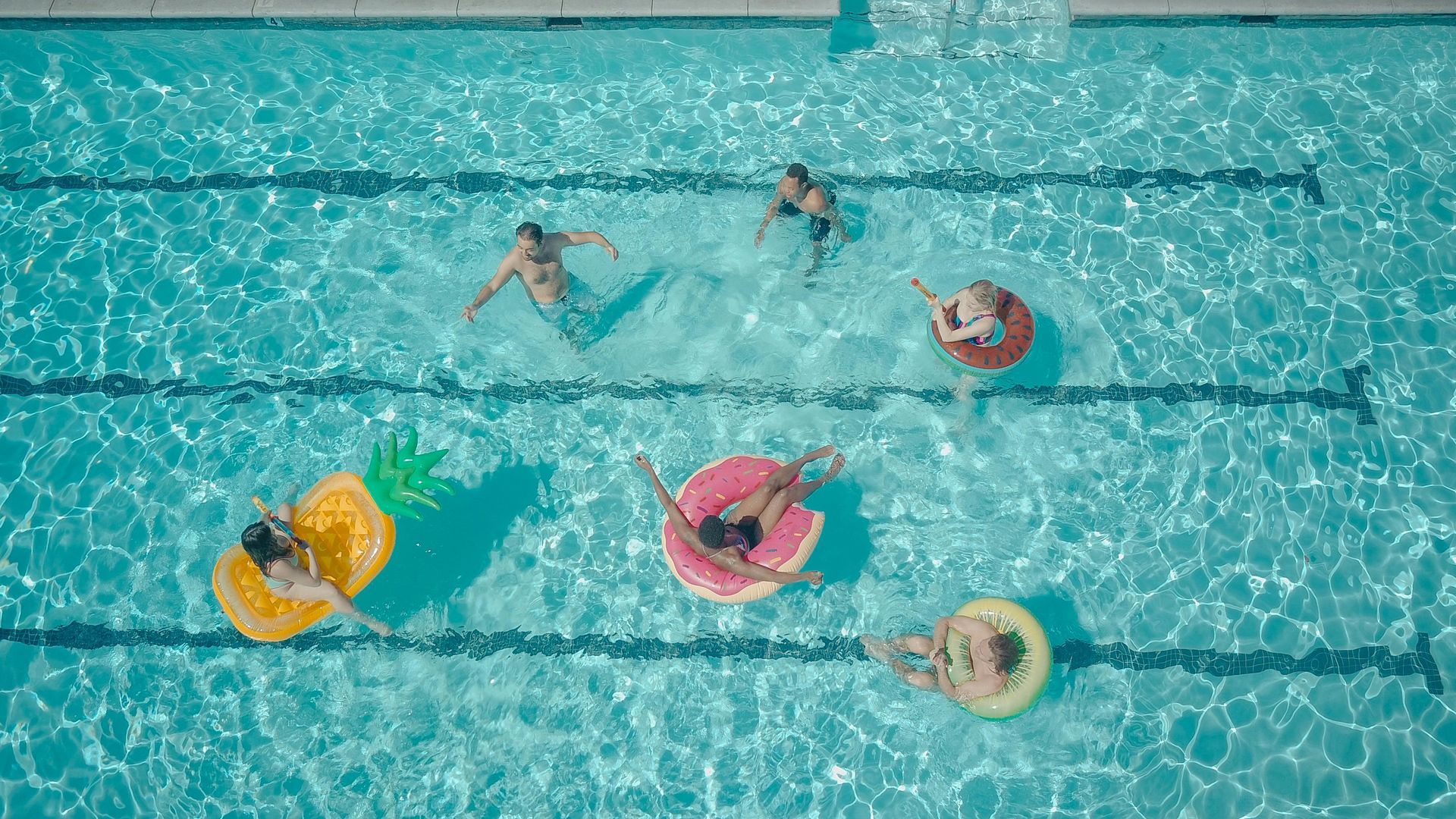 People in a pool with inflatable floats, sunbathing and swimming. Blue water, sunny day.