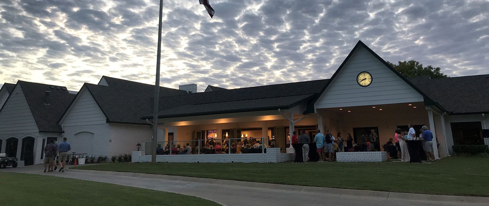 Clubhouse with people gathered on the porch and cyclists on a path, under a cloudy evening sky.