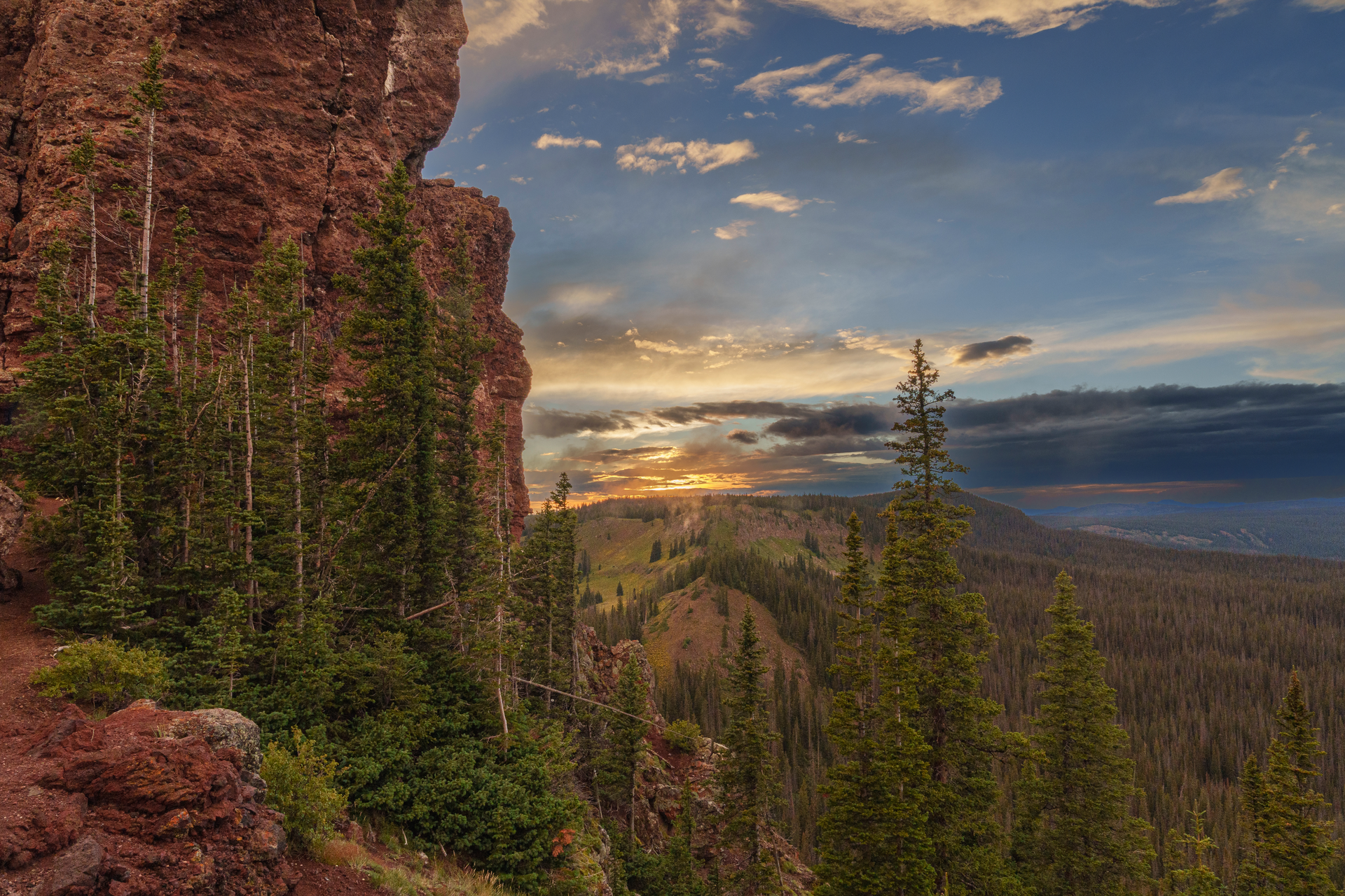 A sunset over a mountain range with trees and rocks in the foreground.