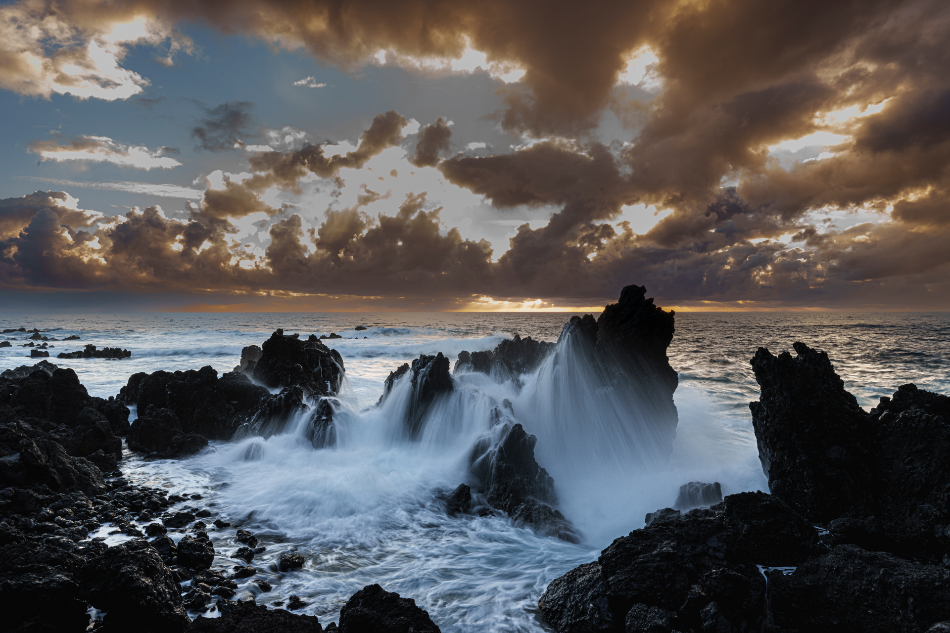 A sunset over the ocean with waves crashing against the rocks