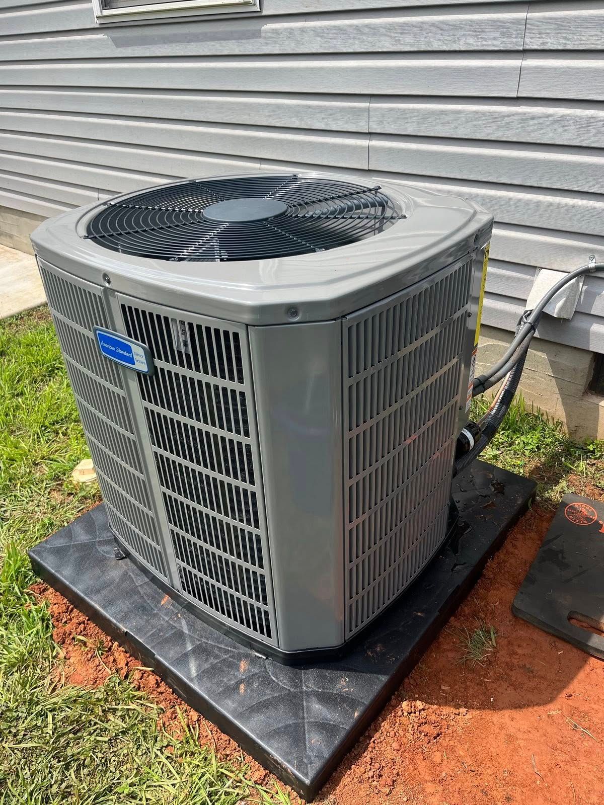A gray air conditioner is sitting on top of a black tray on the side of a house.