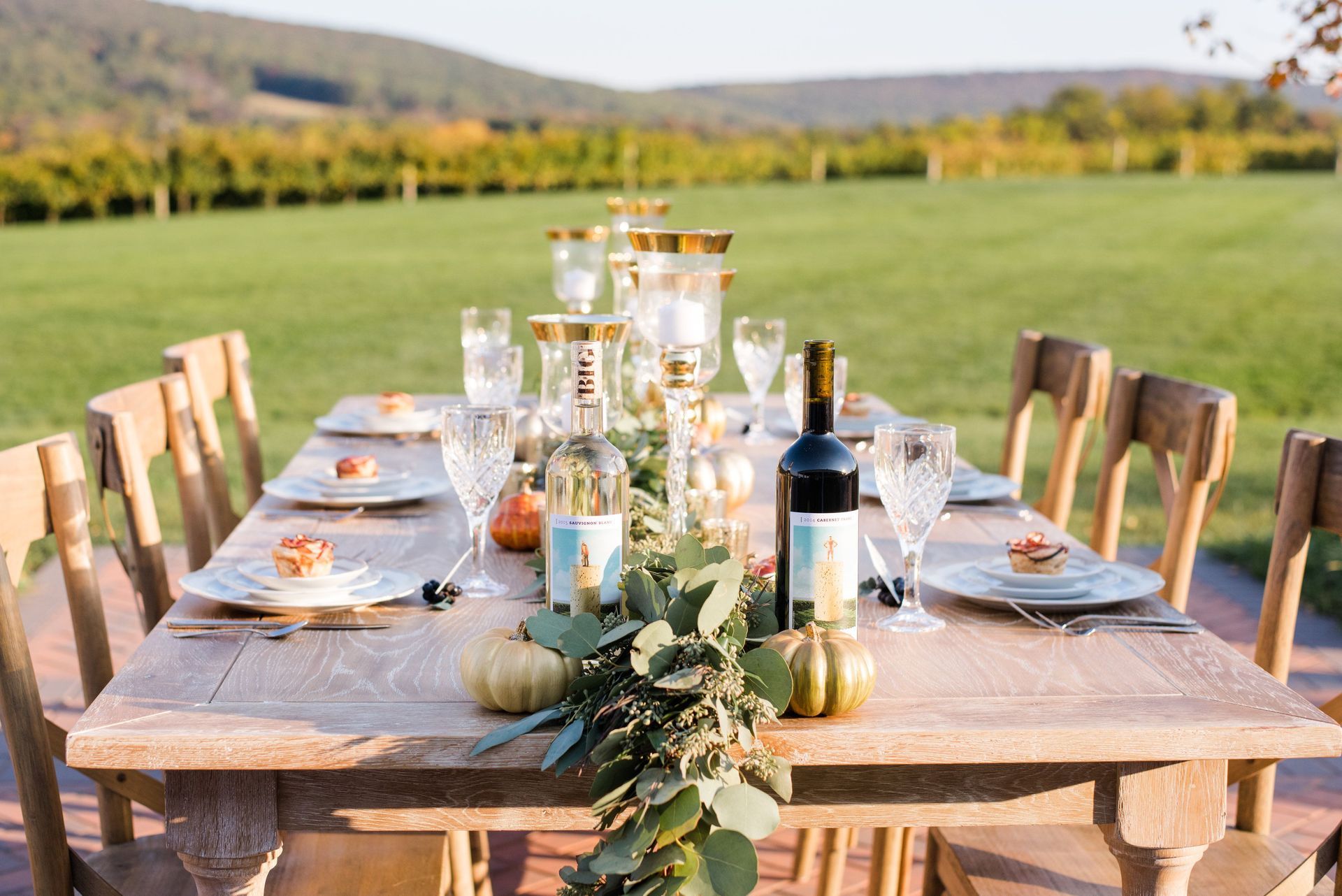 A long wooden table with plates , glasses , and a bottle of wine on it.
