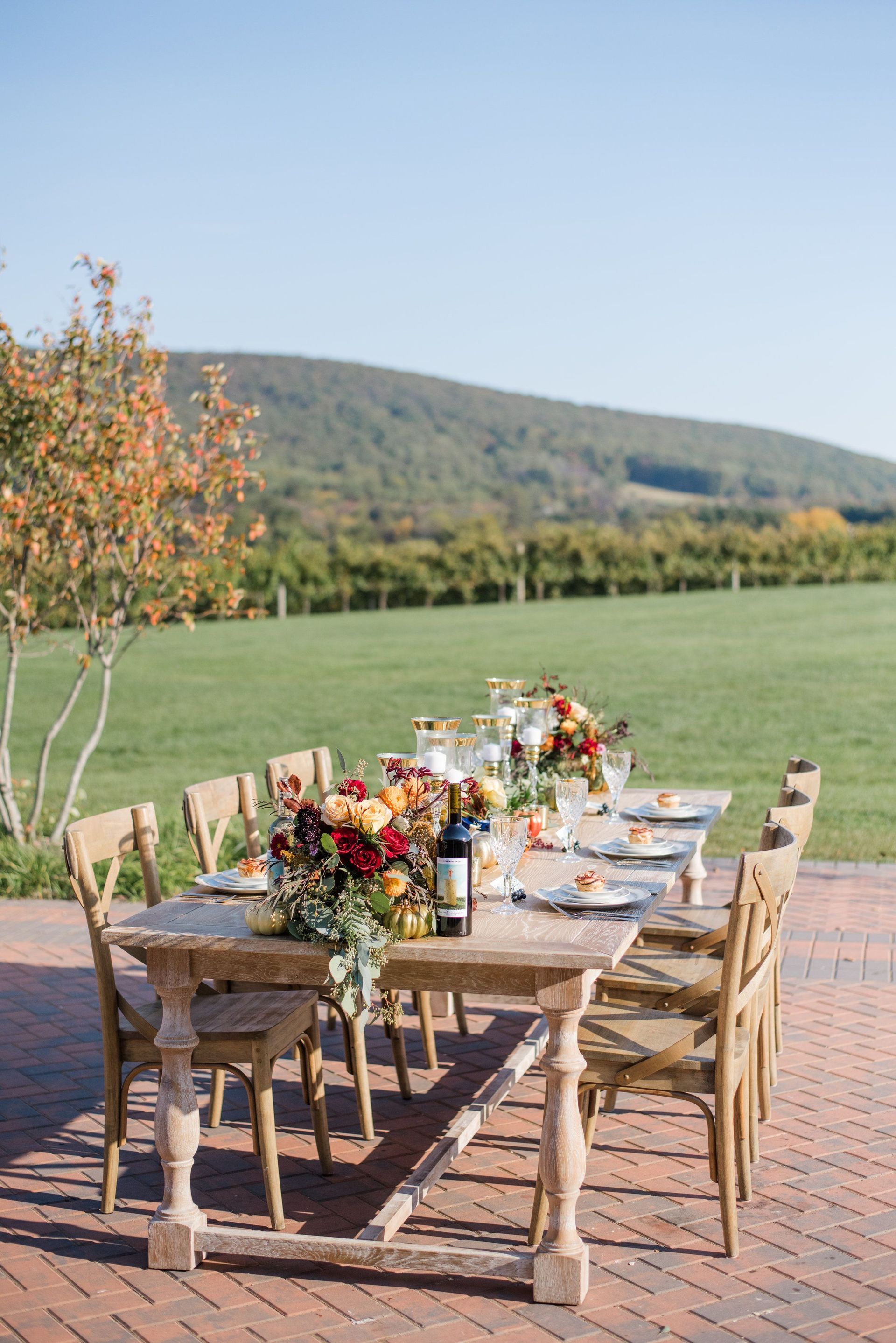 A long wooden table with a bottle of wine on it is sitting on a brick patio.