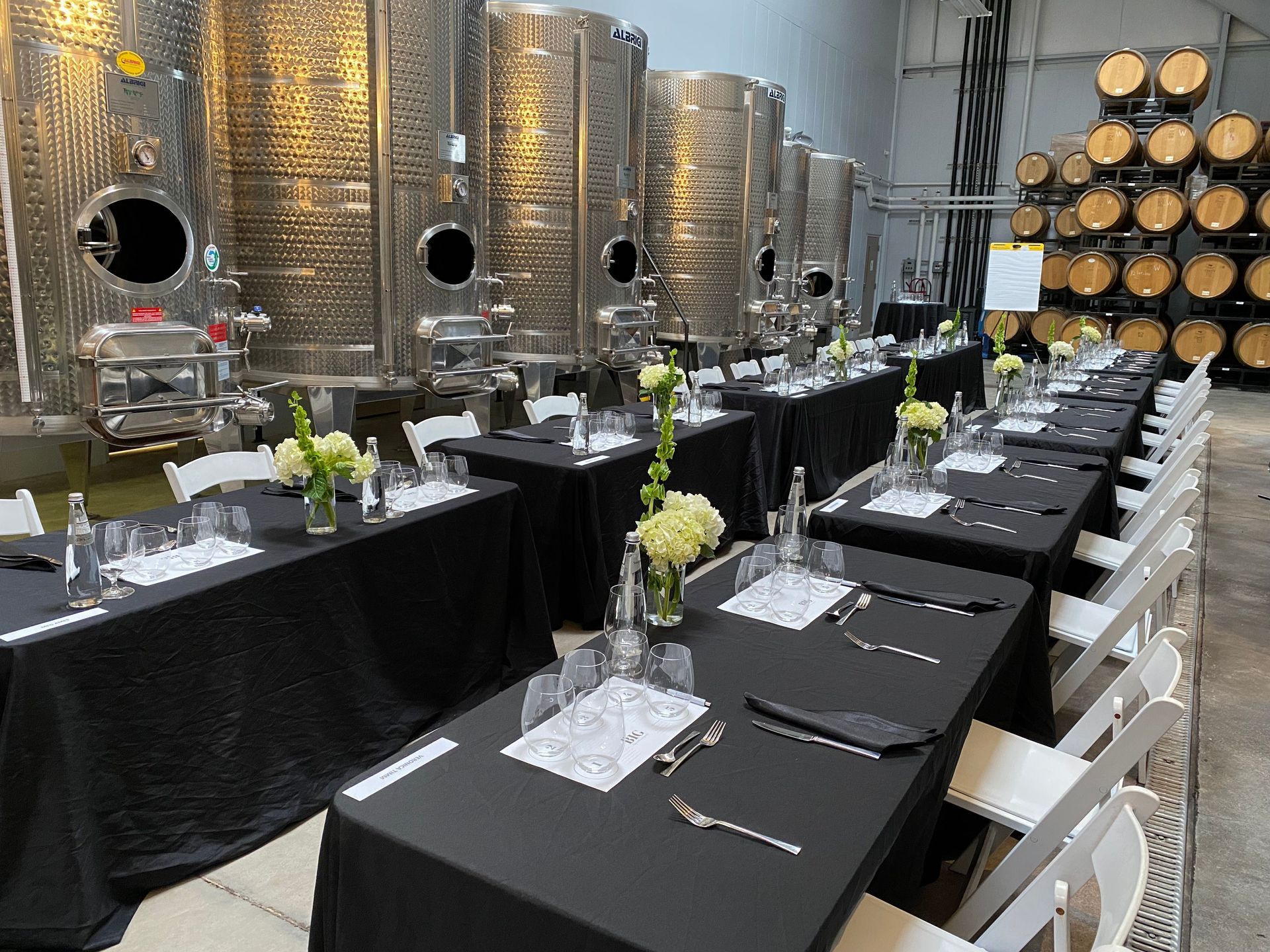 A long row of tables and chairs in a room with barrels in the background.