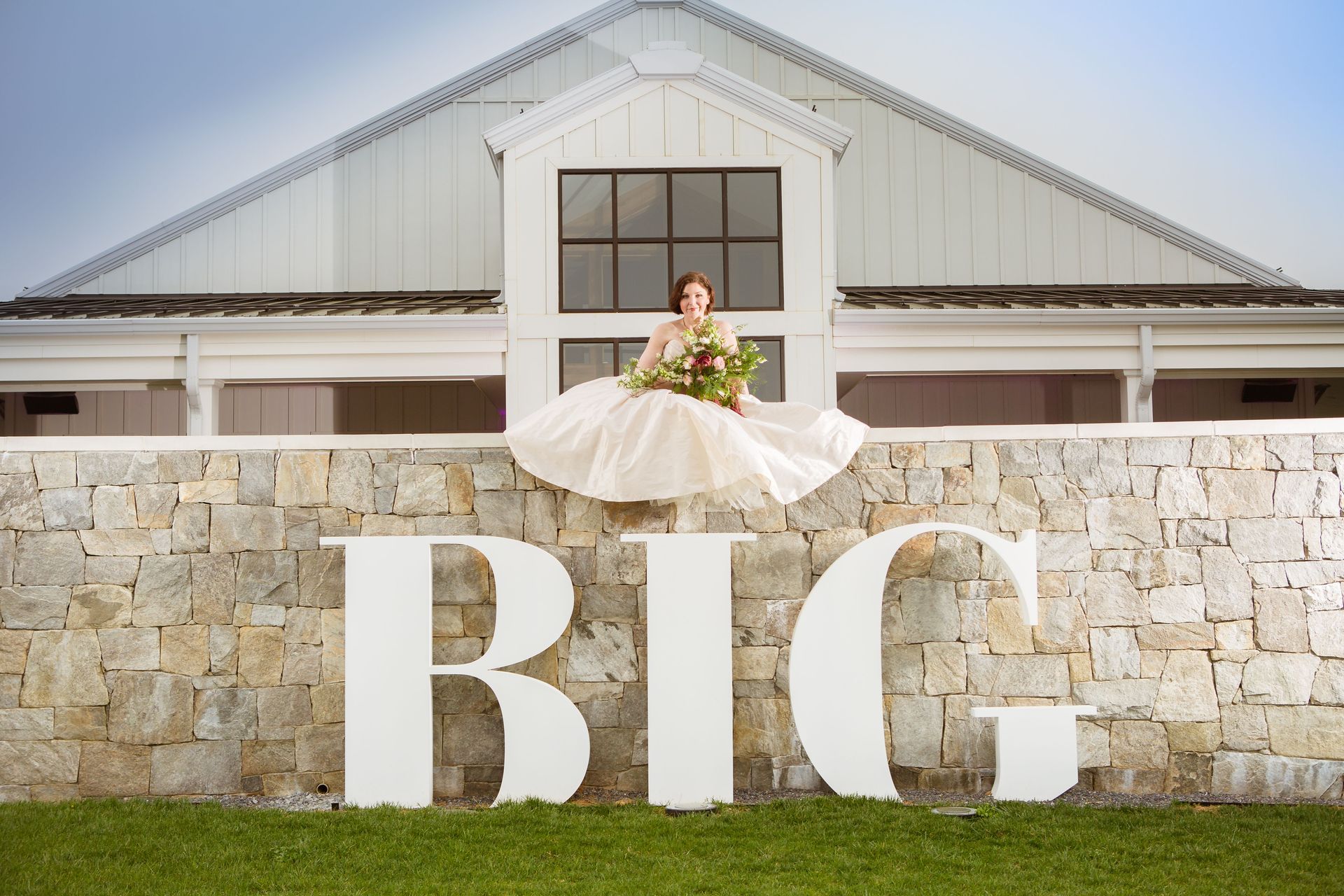 A bride in a wedding dress is sitting on a stone wall in front of a white building.