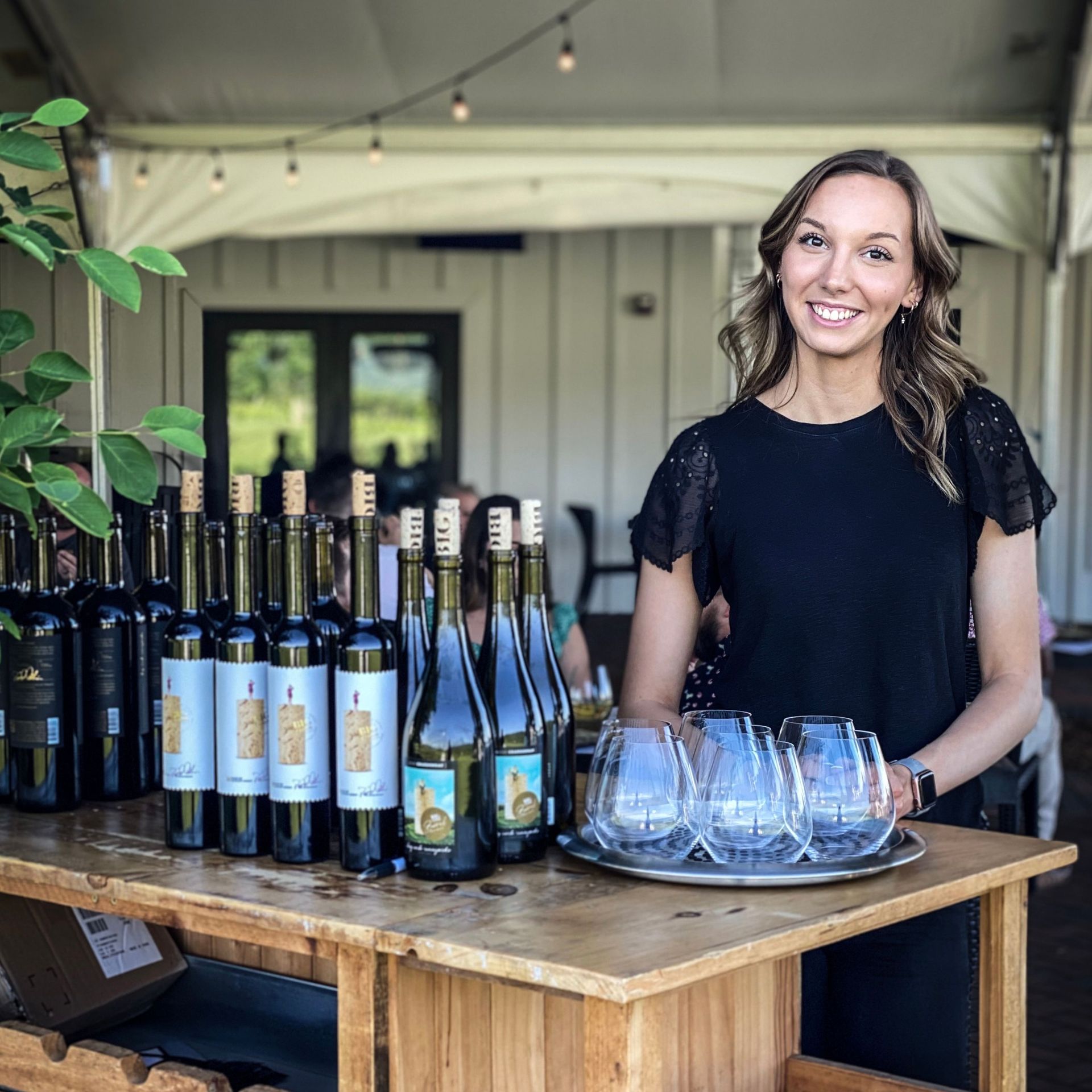 A woman is standing in front of a table full of wine bottles and glasses.