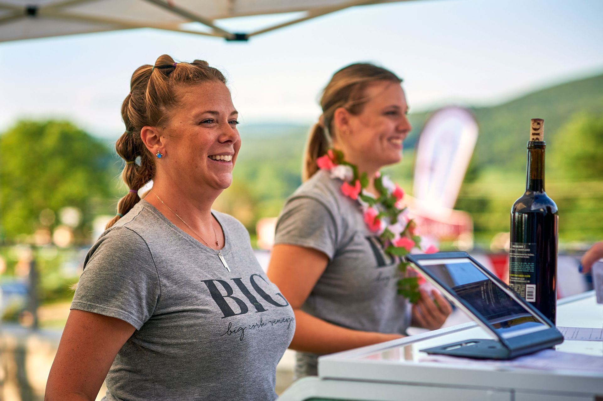 Two women are standing next to each other at a table with a laptop and a bottle of wine.