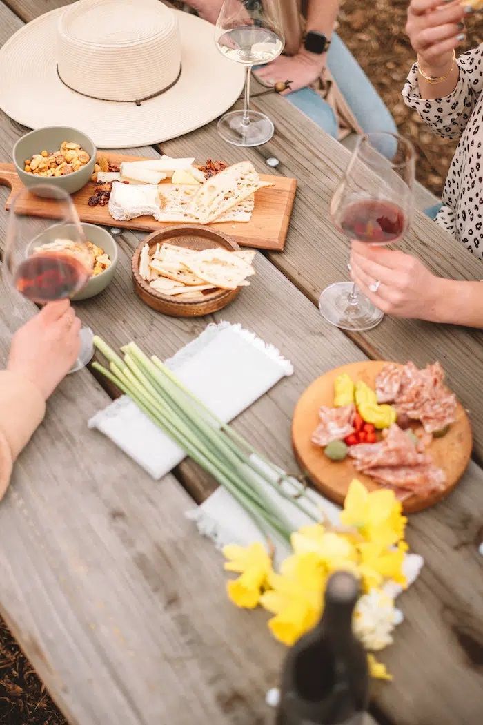 A group of people are sitting at a picnic table eating food and drinking wine.