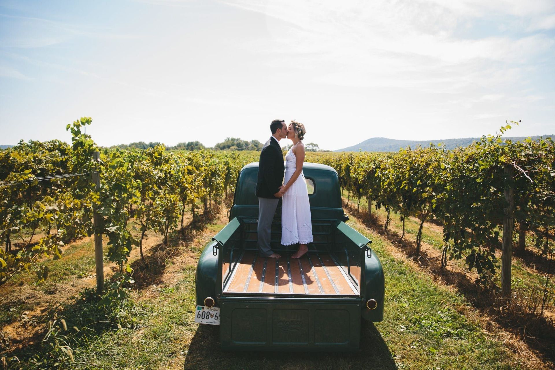 A bride and groom are kissing in the back of a truck in a vineyard.