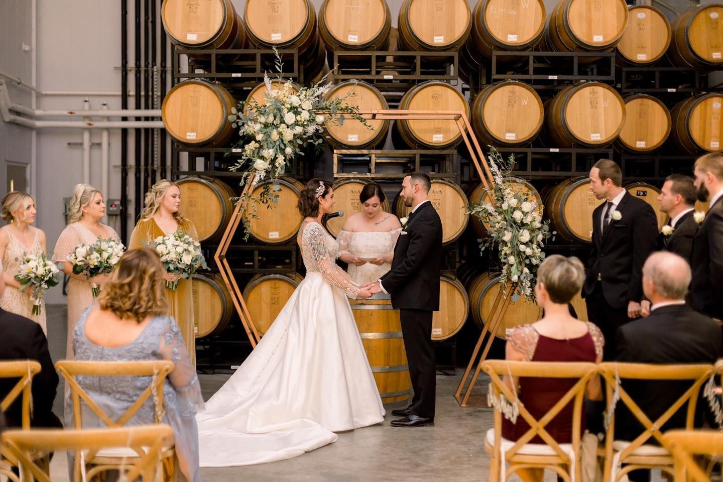 A bride and groom are holding hands during their wedding ceremony in front of a wall of wine barrels.