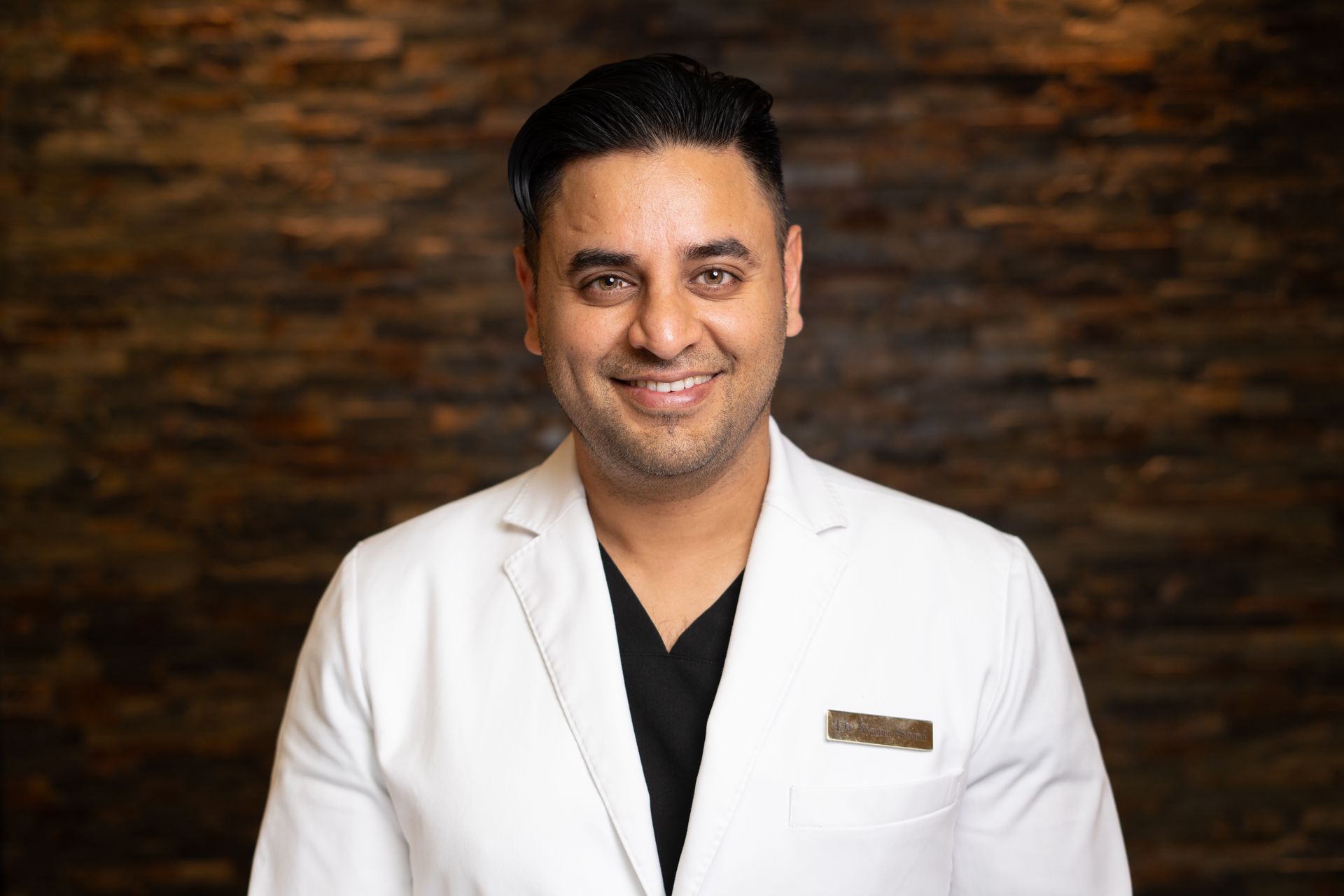 Man in white coat smiles, standing in front of a textured brown wall.