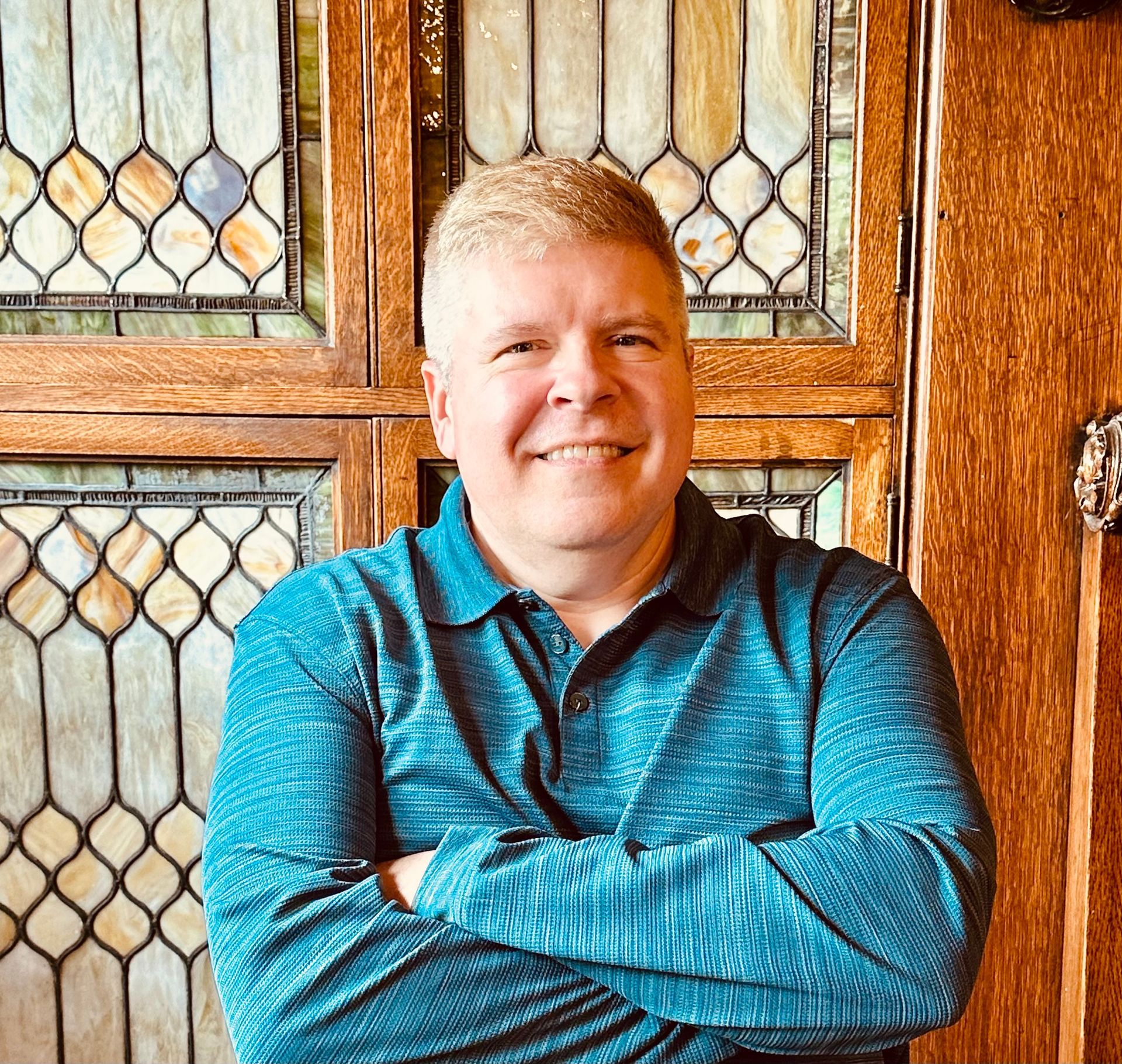 A man in a blue shirt is standing with his arms crossed in front of a stained glass window.
