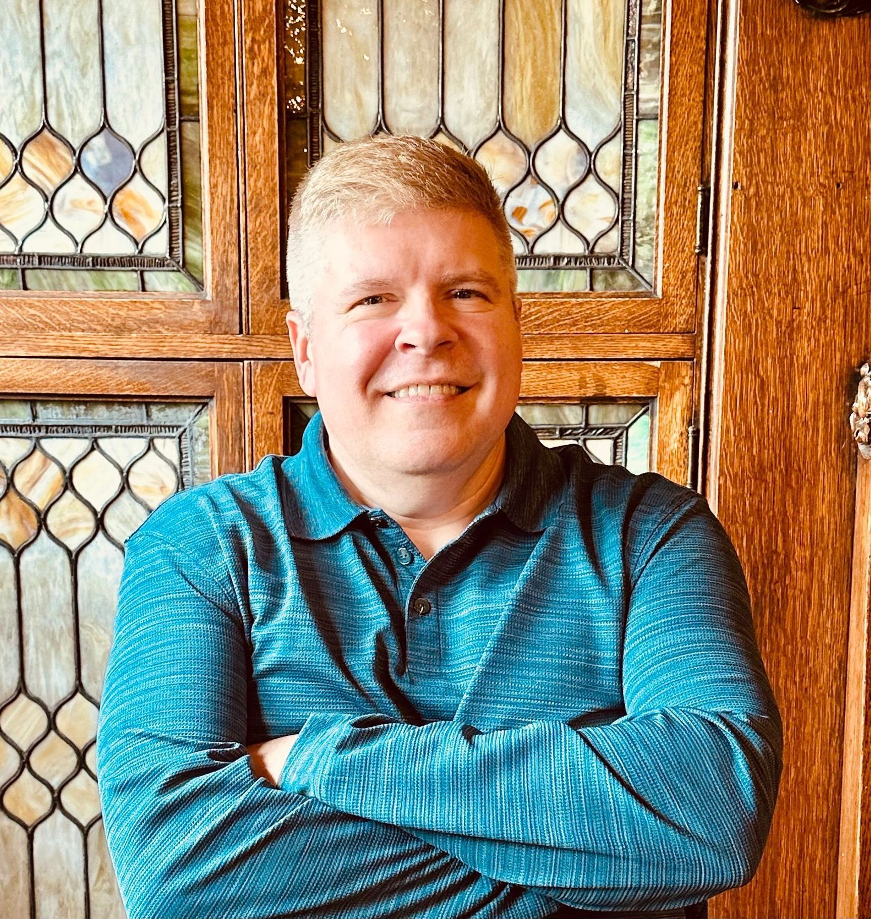 A man in a blue shirt is standing with his arms crossed in front of a stained glass window.