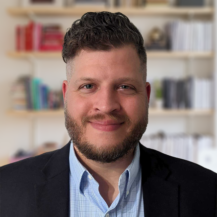 A man with a beard and a suit is smiling in front of a bookshelf.