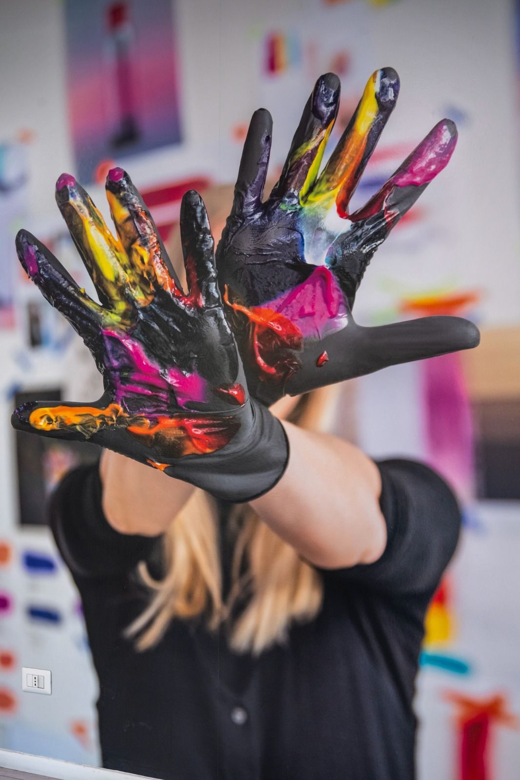 Persona che alza le mani ricoperte di vernice colorata, di fronte allo sfondo colorato di uno studio d'arte.
