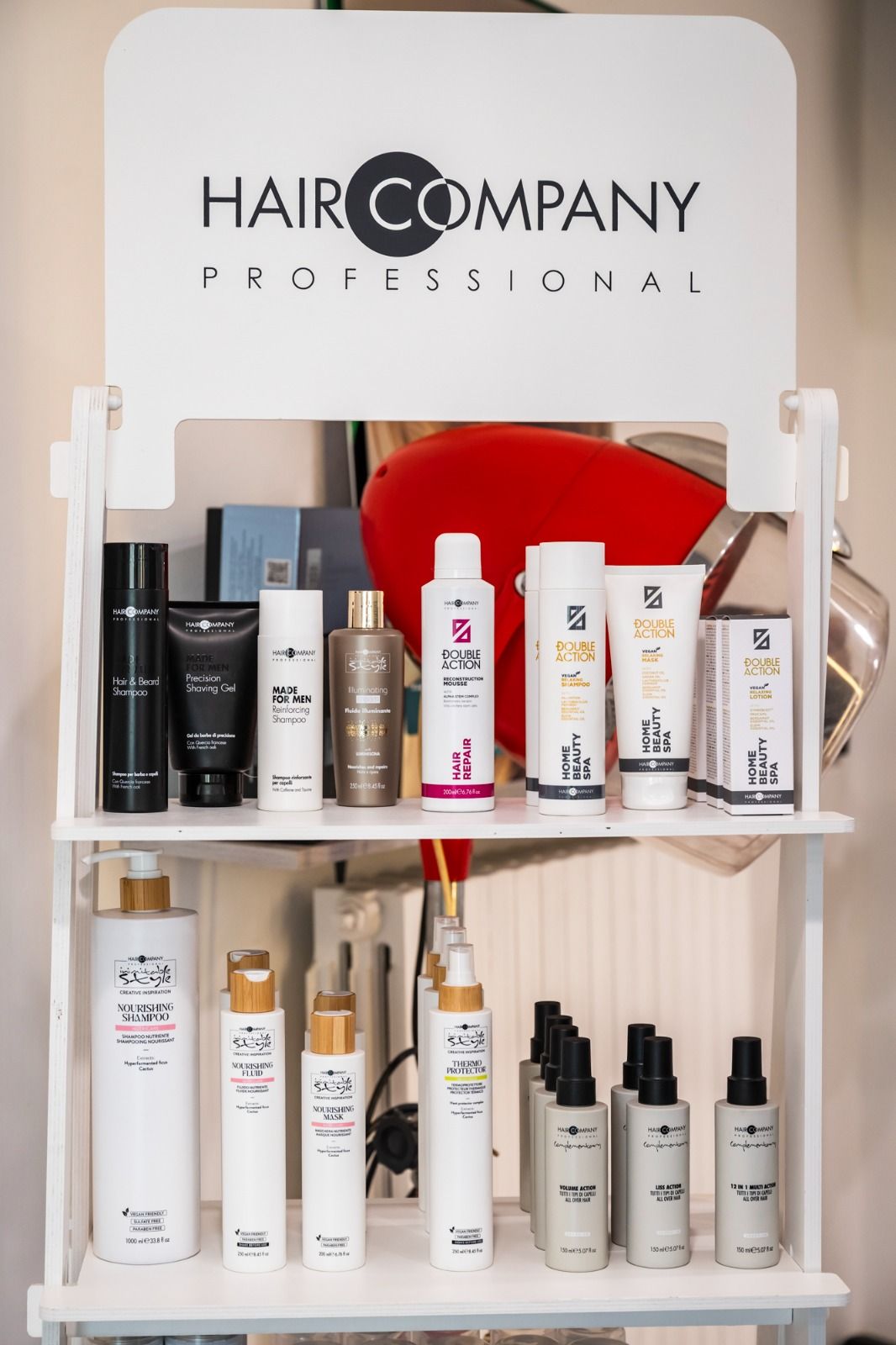 Espositore con prodotti per capelli Hair Company Professional, ripiani bianchi, vari flaconi, parete beige.