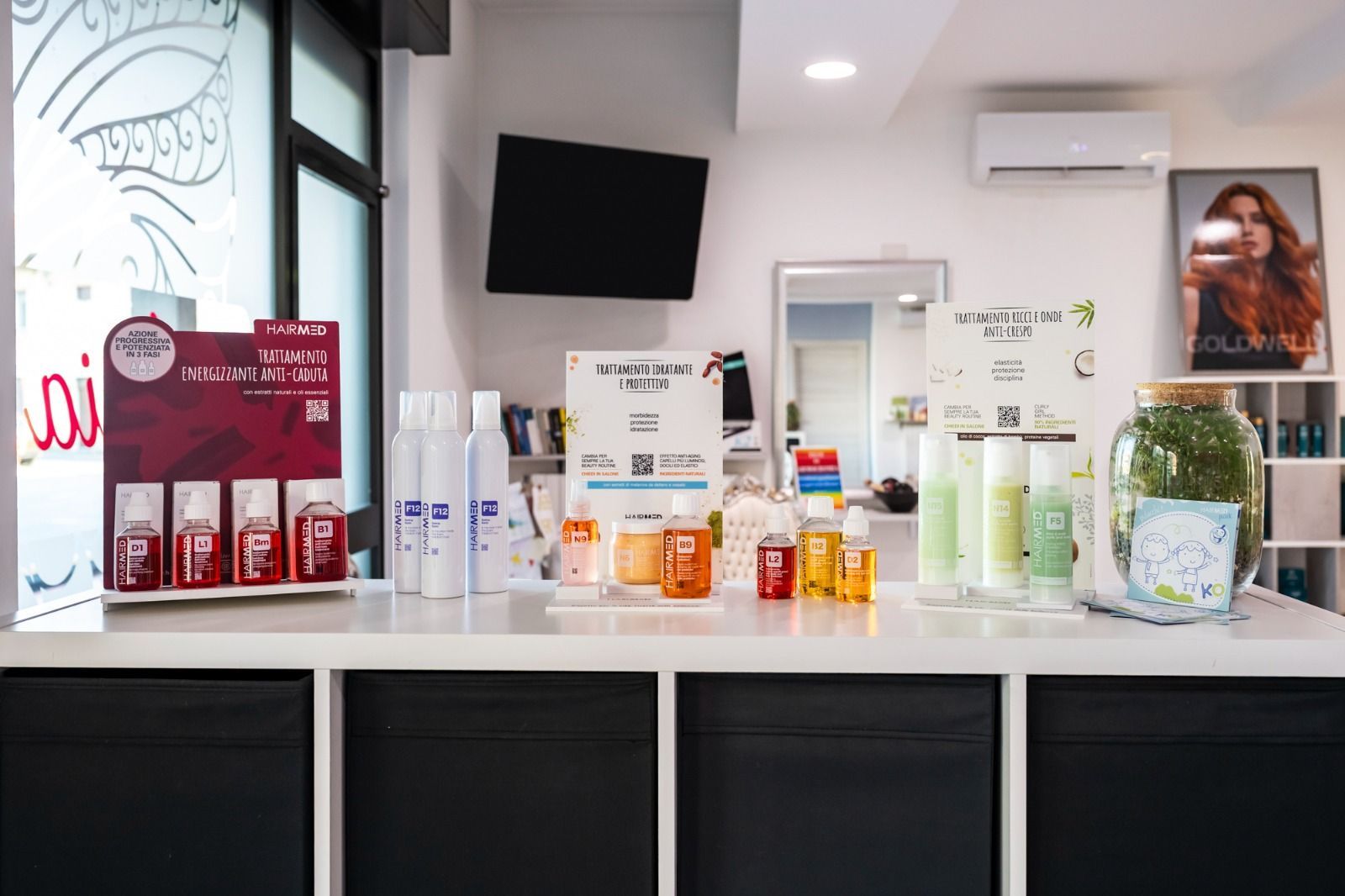 Prodotti per capelli esposti su un bancone bianco in un salone, tra cui diverse bottiglie e un grande barattolo con del verde.