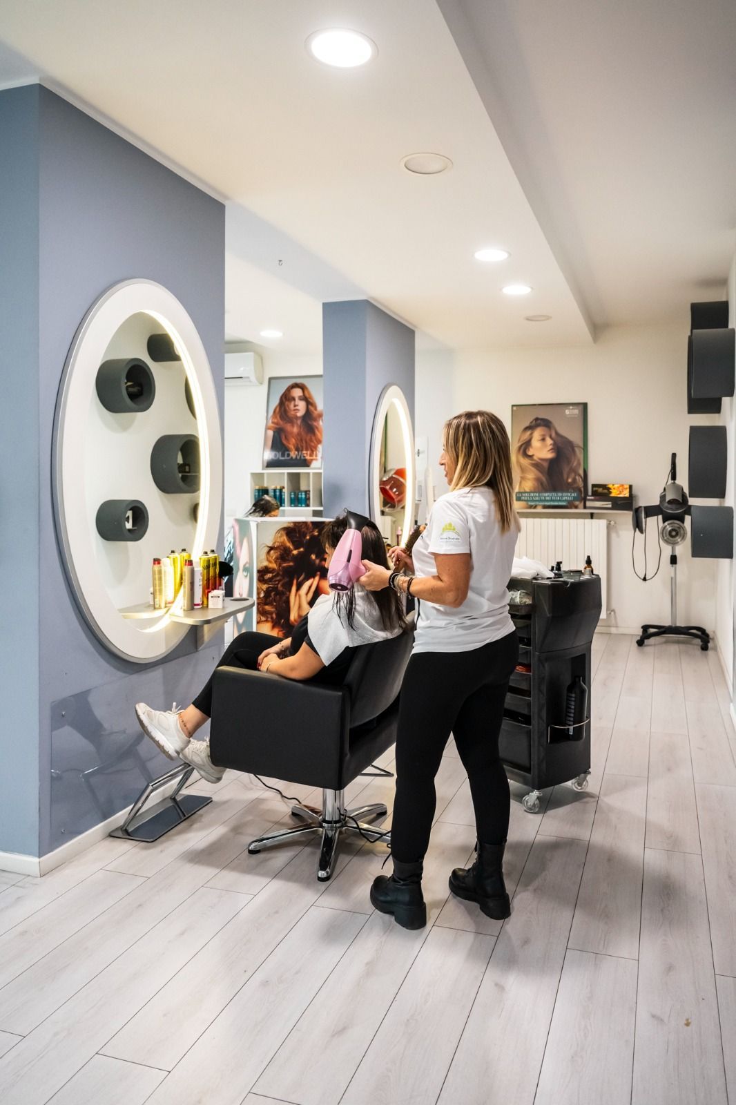 Parrucchiere che acconcia i capelli di una cliente in un salone. La cliente è seduta, mentre il parrucchiere è in piedi con un asciugacapelli.