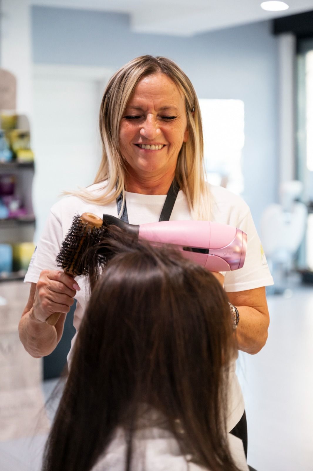 Parrucchiere che usa una spazzola e un asciugacapelli rosa sui capelli scuri di una cliente in un salone; sorride.