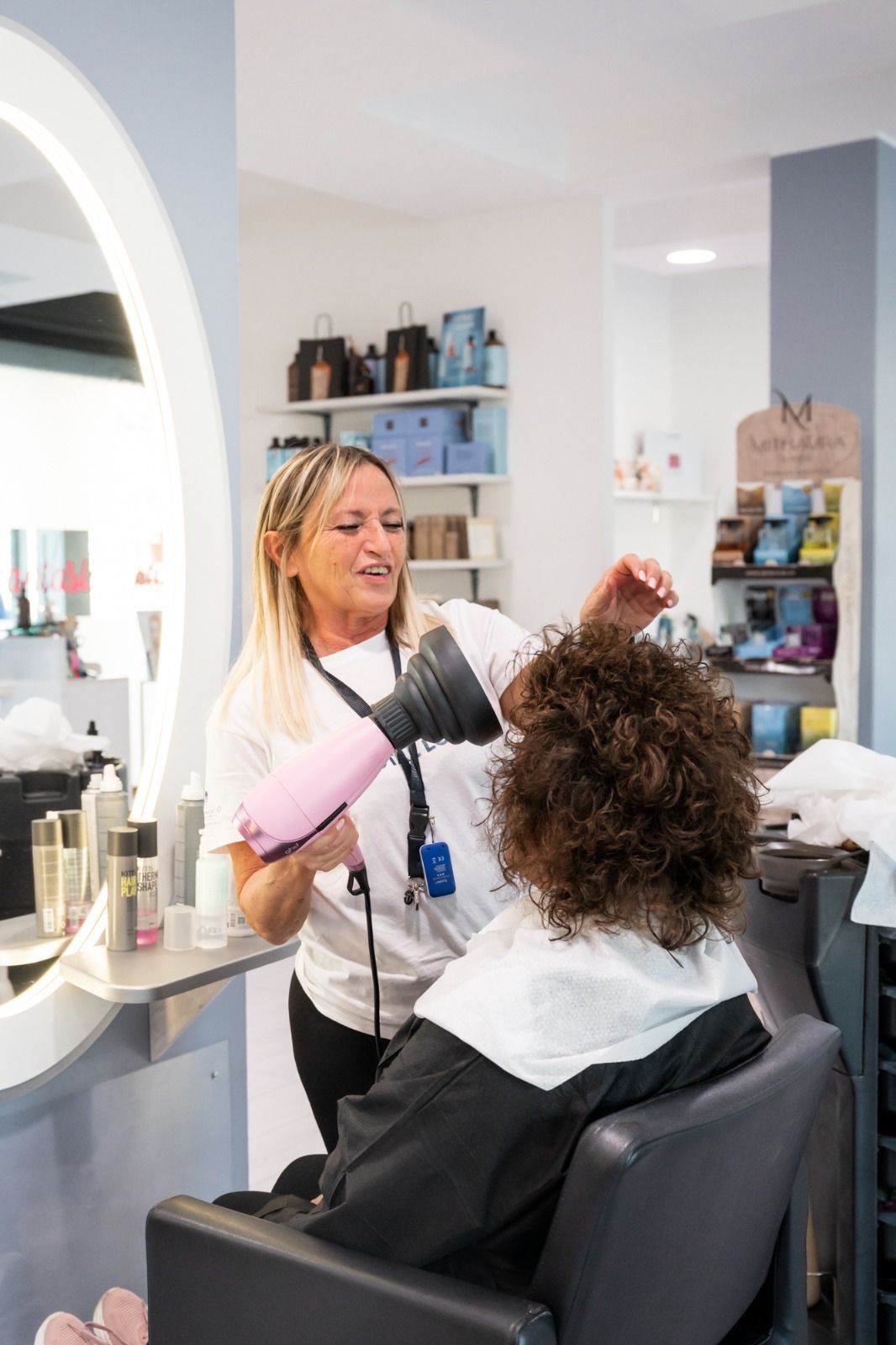 Parrucchiere che usa un asciugacapelli rosa sui capelli ricci di una cliente in un salone con pareti bianche e un grande specchio.