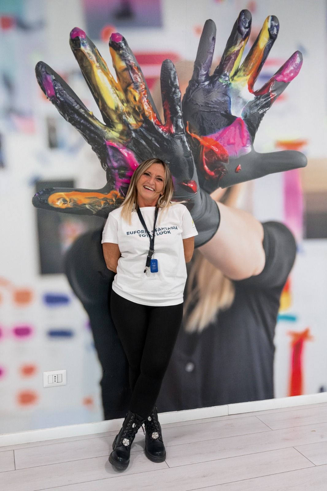 Una donna con una maglietta bianca e pantaloni neri è in piedi davanti a un murale colorato con l'impronta di una mano.