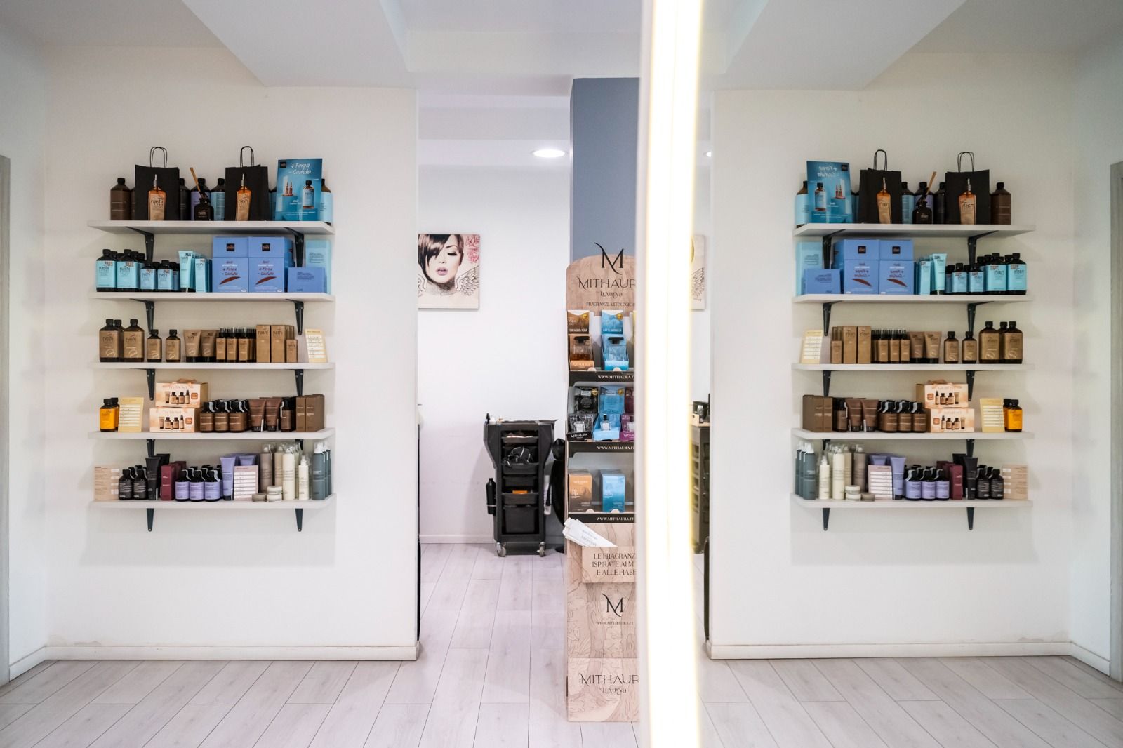 Interno di un salone da parrucchiere con scaffali pieni di prodotti su pareti bianche, un lungo specchio e una lampada.